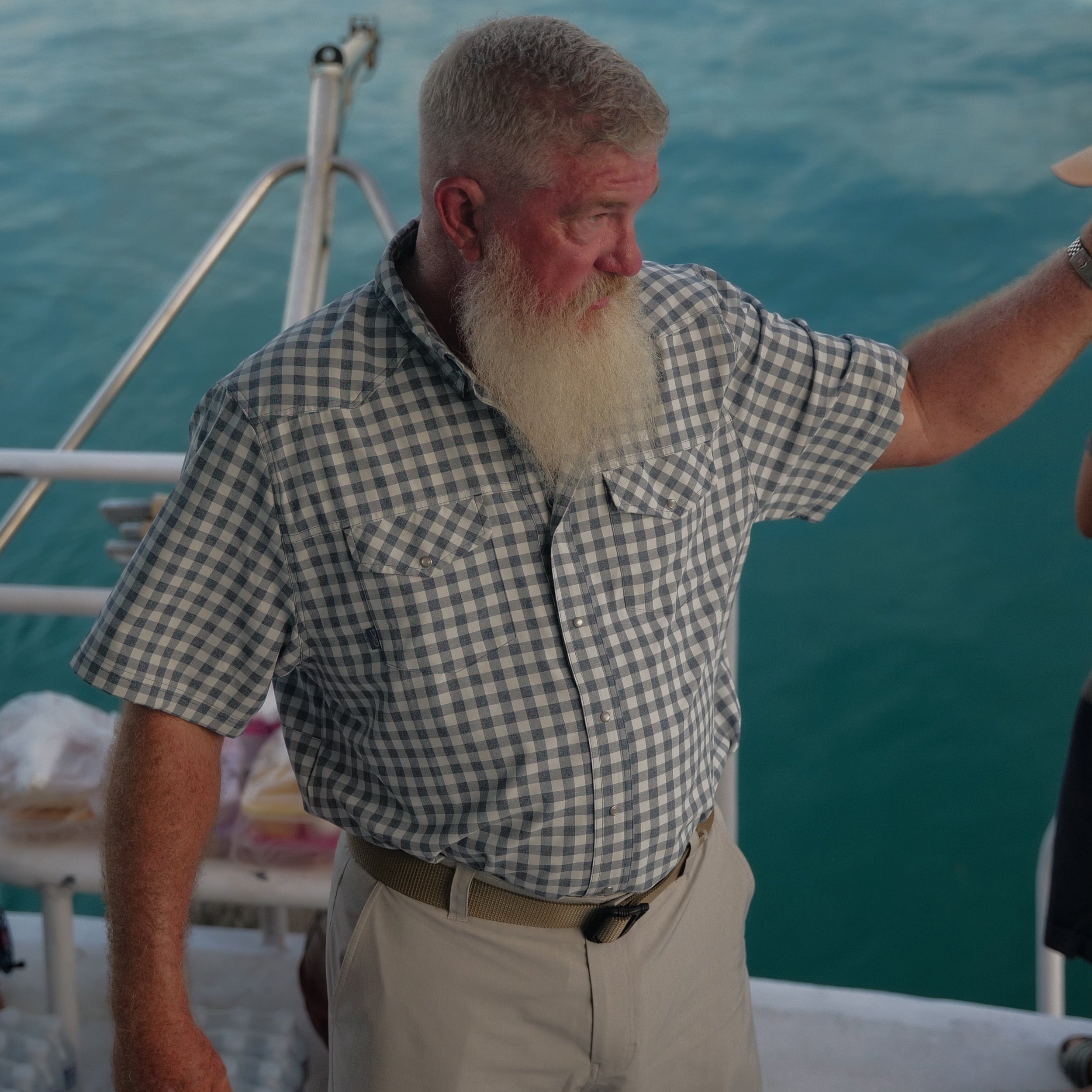 Man on a boat wearing a checkered shirt with a blurred background of water and deck.