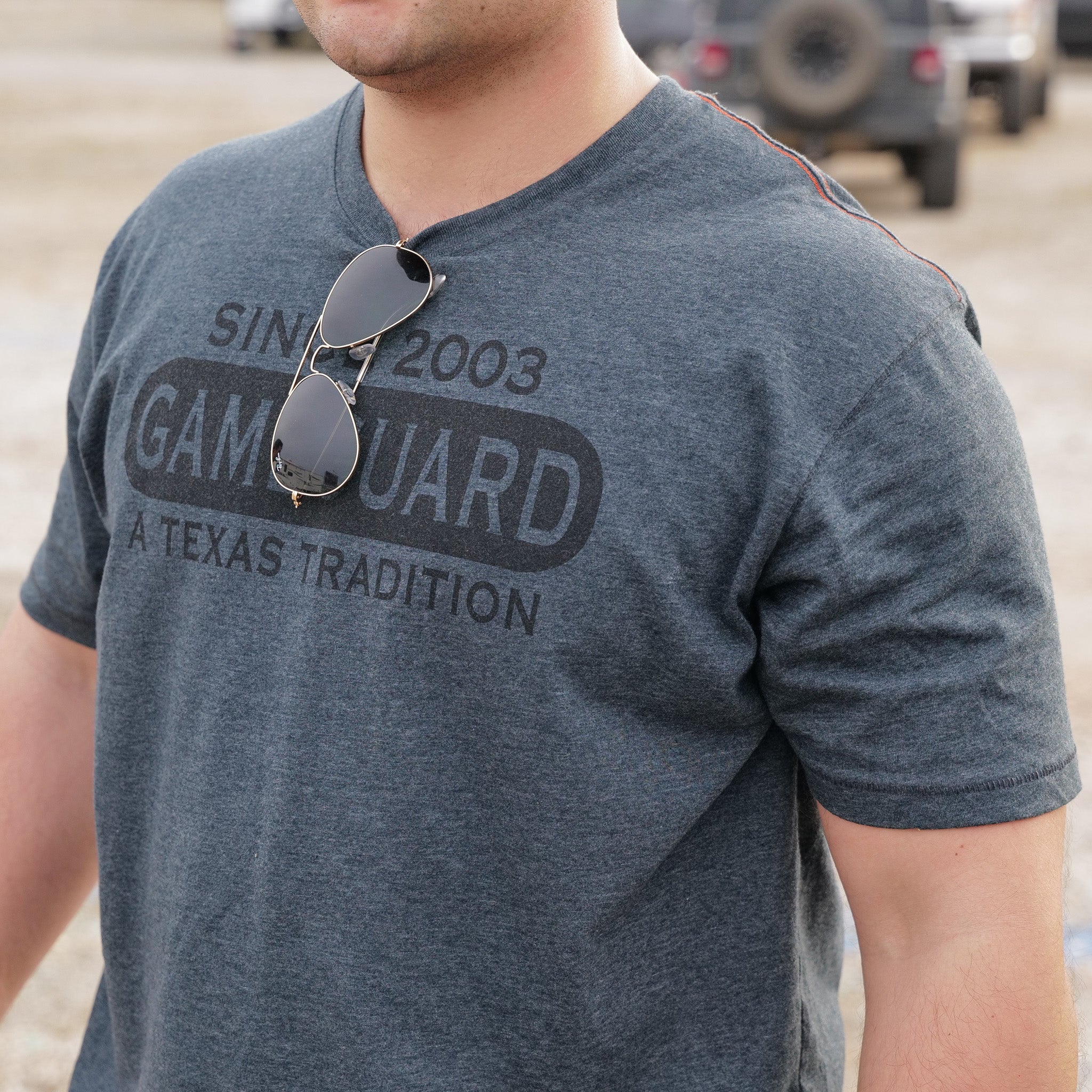 Person wearing a gray t-shirt with 'Game Guard' text, sunglasses on shirt, blurred outdoor background