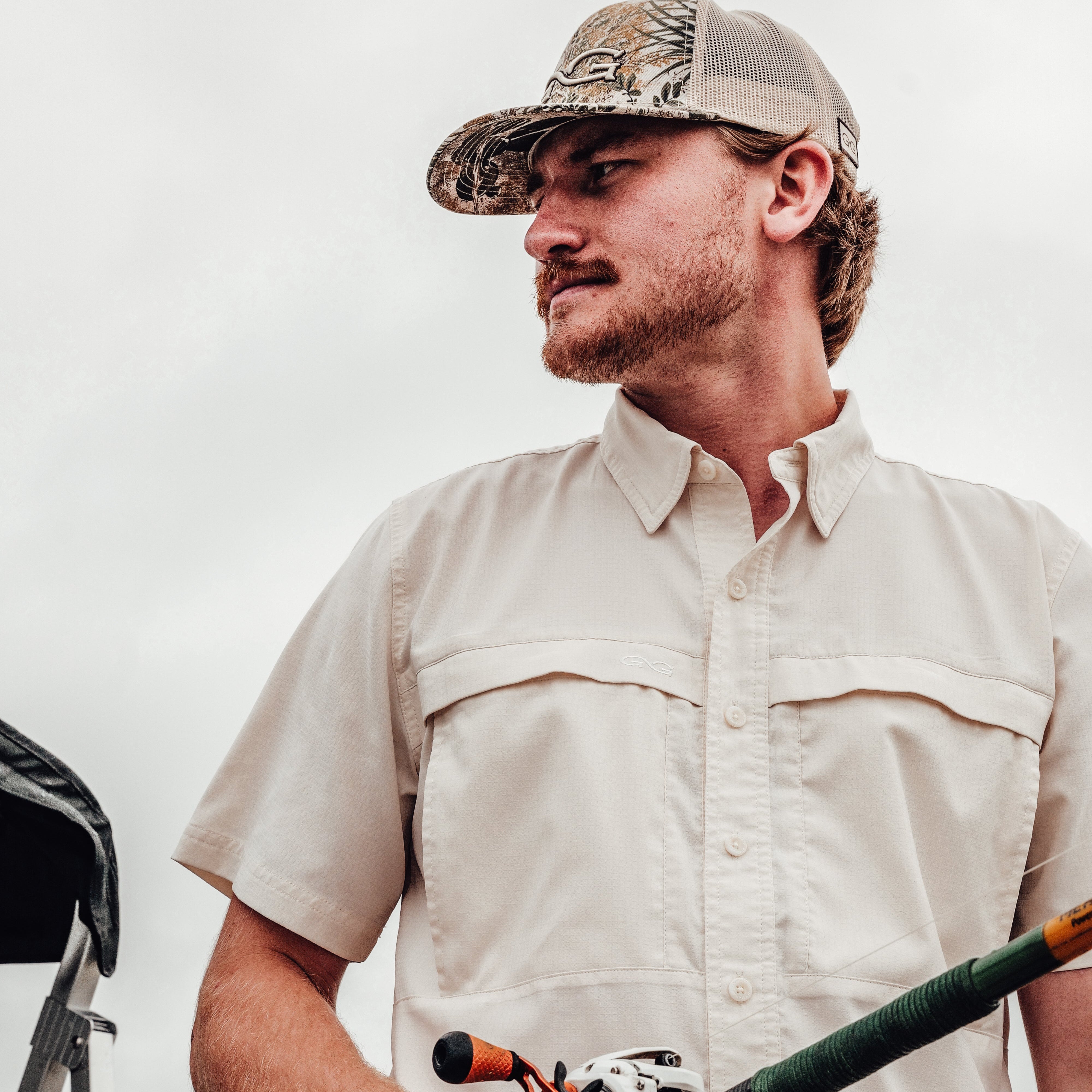 Man wearing a beige shirt and camouflage cap holding a fishing rod against a white background