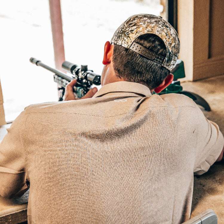 Man in camouflage cap and beige jacket aiming a rifle with a scope.