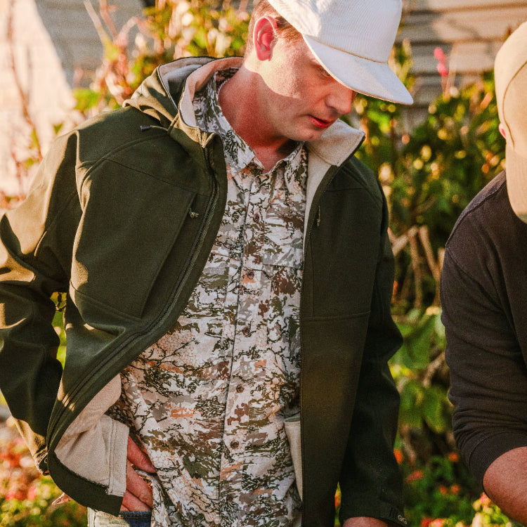 Person wearing a green jacket and camouflage shirt with a white cap, standing outdoors.