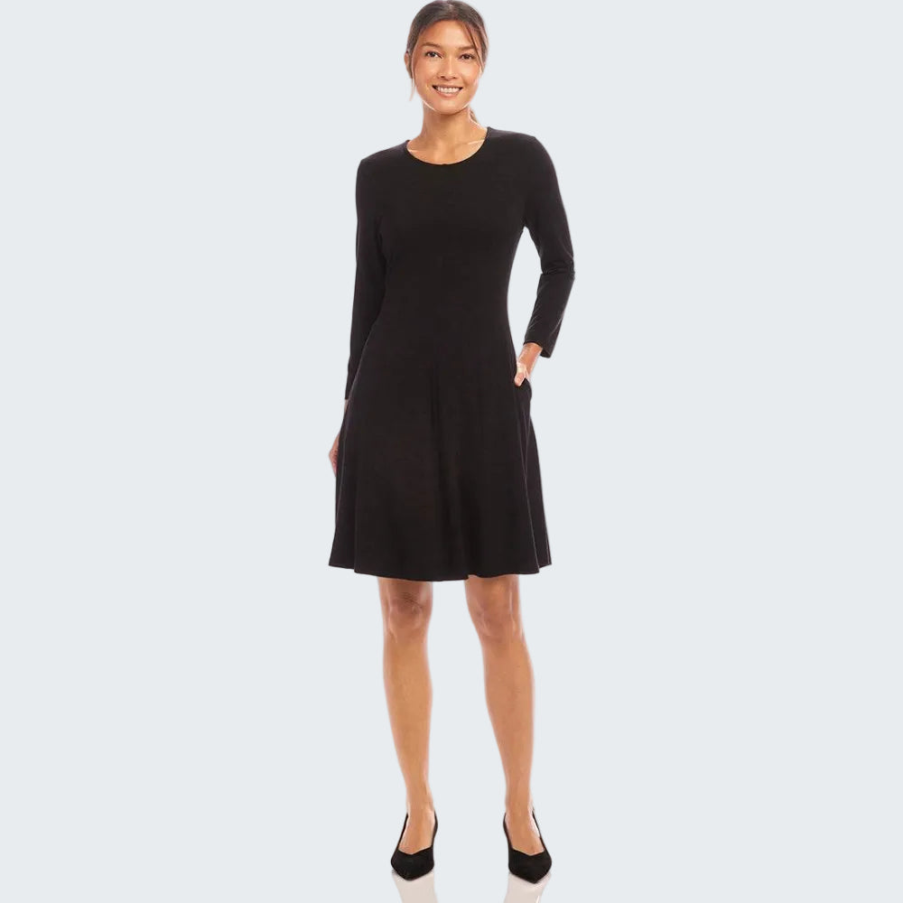A woman modeling a black long-sleeve shift dress with a round neckline and side pockets, paired with black pointed-toe heels, against a plain white studio background.