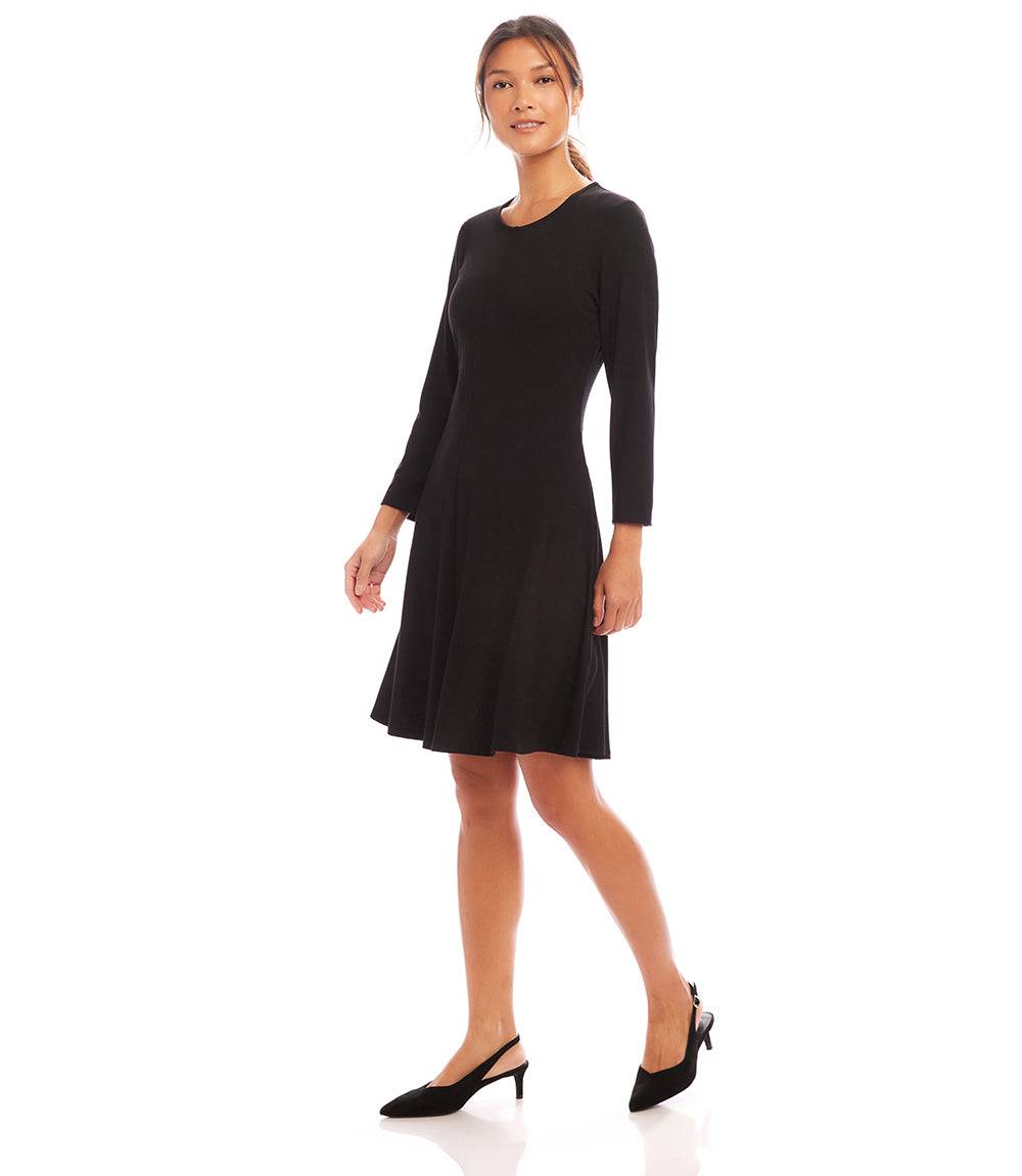 A woman modeling a black long-sleeve knee-length shift dress with a flared skirt, paired with black slingback heels, against a plain white studio background.