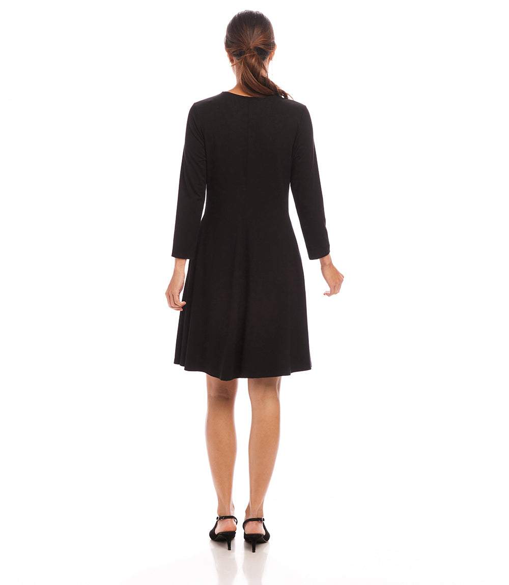 A woman viewed from the back wearing a black long-sleeve knee-length dress and black heels against a plain white studio background.
