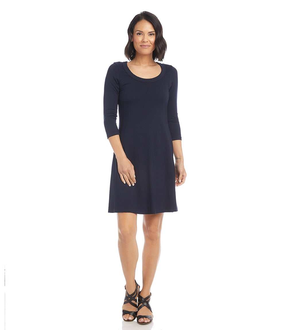 A woman modeling a dark navy blue three-quarter sleeve shift dress with a scoop neckline, paired with black strappy sandals, against a plain white background.
