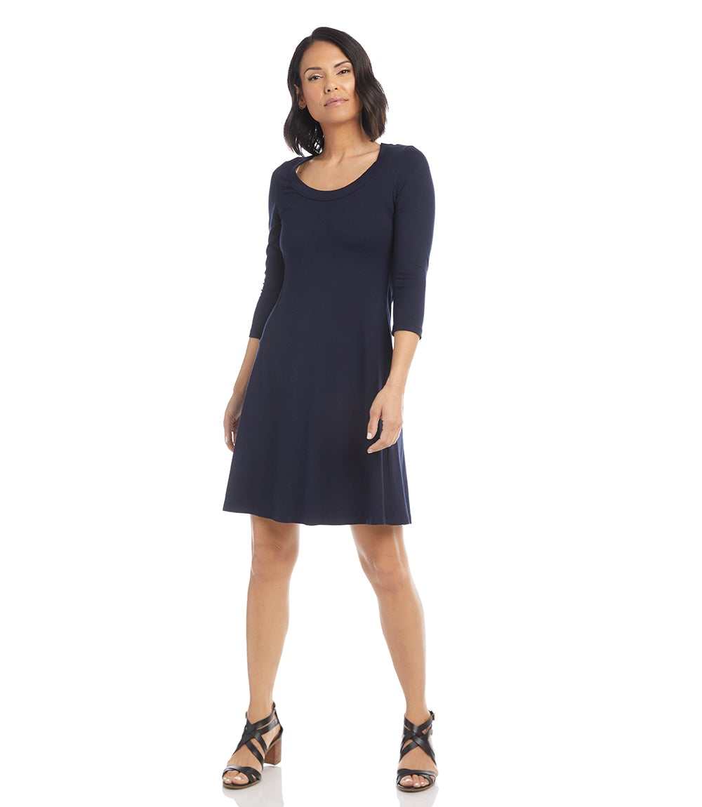 A woman modeling a navy blue three-quarter sleeve shift dress with a scoop neckline, paired with black strappy sandals, against a solid white background.