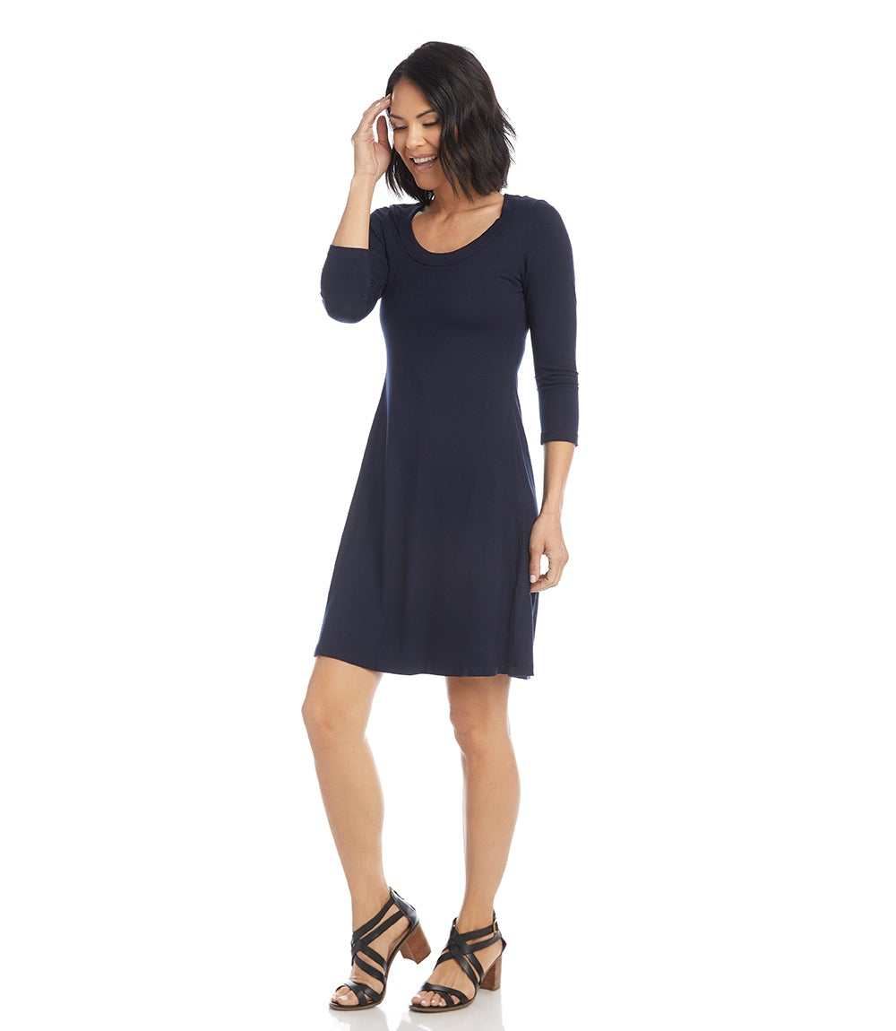 A woman modeling a dark navy blue three-quarter sleeve shift dress with a scoop neckline, paired with black strappy high-heeled sandals, against a plain white background.
