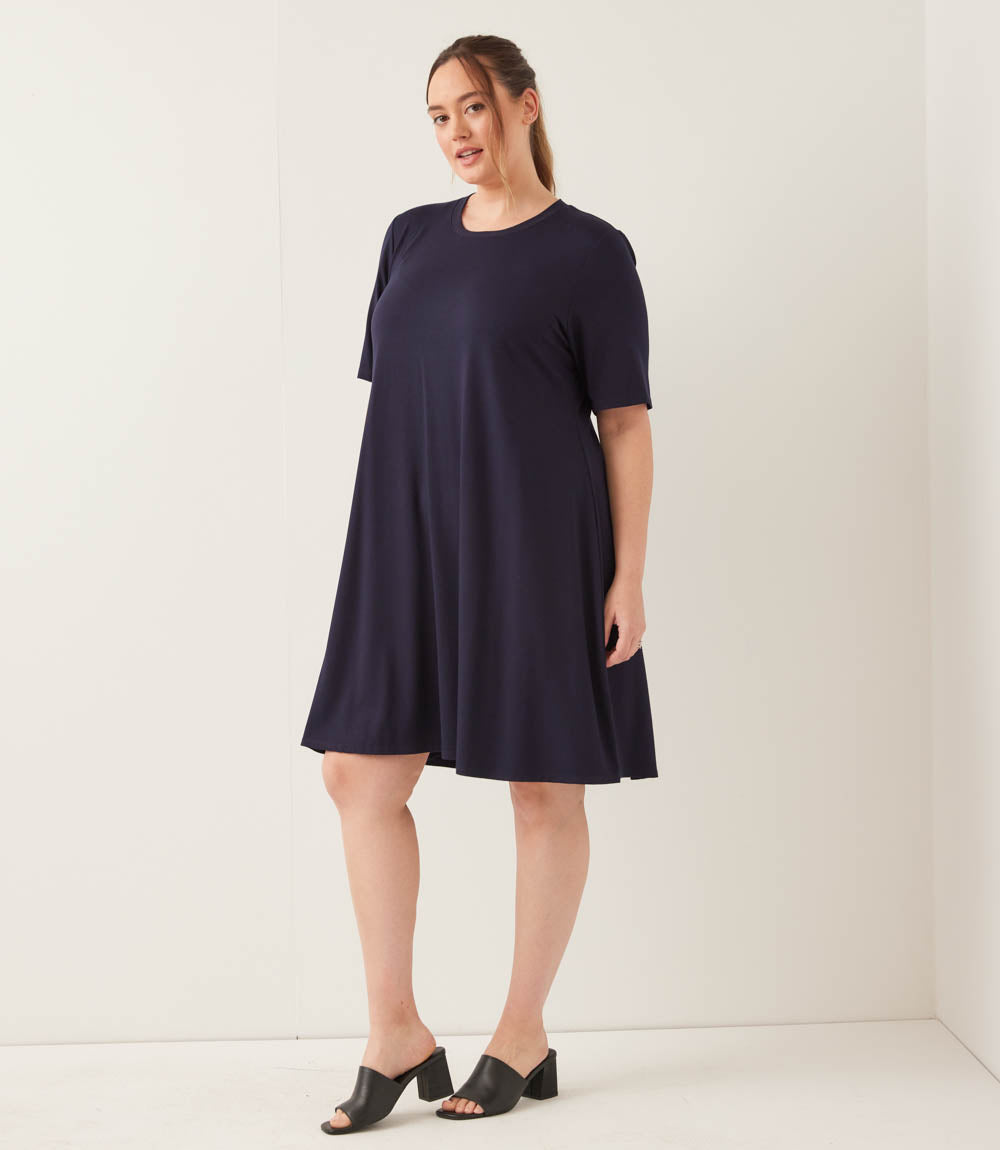 A woman modeling a short-sleeve, knee-length navy blue shift dress with black mule sandals, standing against a plain off-white studio background.