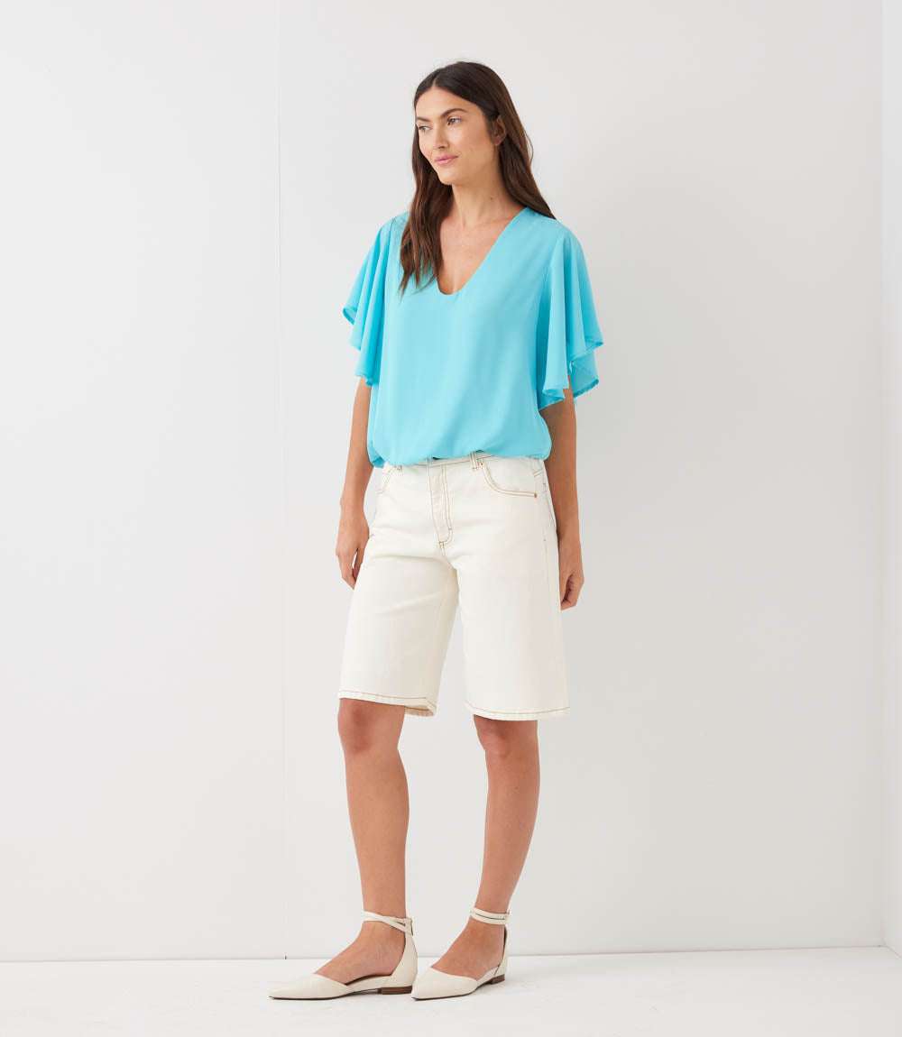 A woman modeling a light blue short-sleeve V-neck blouse with flutter sleeves, paired with off-white denim shorts and white flat sandals, standing against a plain white studio background.