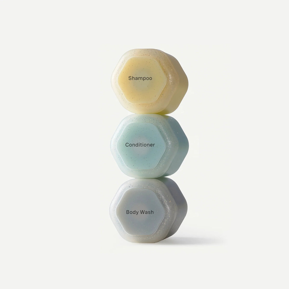 Three hexagonal shampoo and conditioner bars stacked vertically on a clean white background. The top bar is a pale yellow and labeled 'Shampoo', the middle bar is a pale blue and labeled 'Conditioner', and the bottom bar is a very light gray and has a small bar graphic.