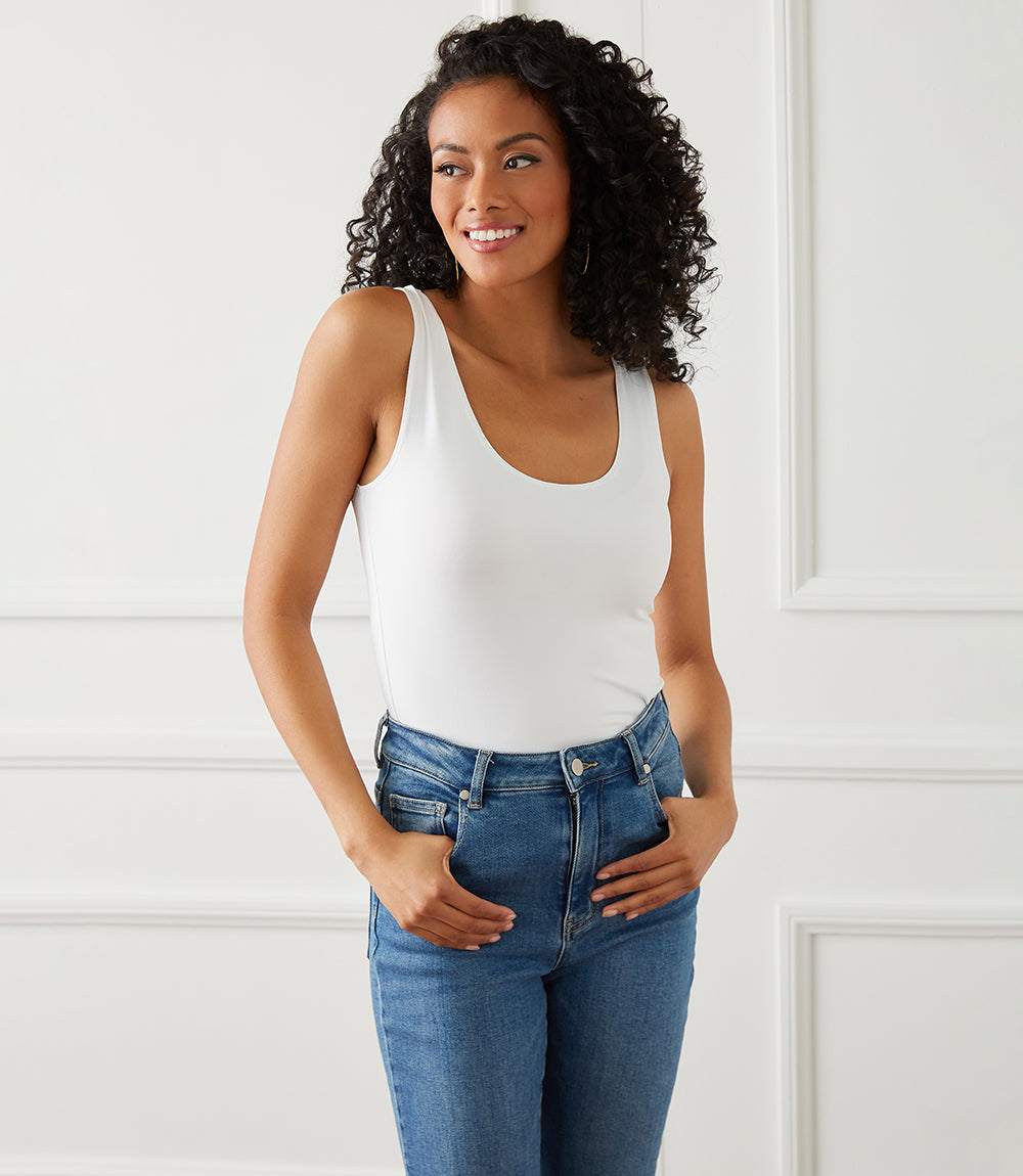 A woman with curly black hair wearing a white scoop-neck tank top and blue jeans, standing against a white paneled wall.