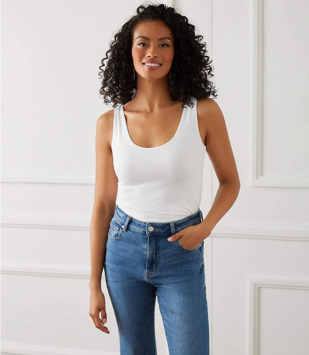 Woman wearing a white tank top and blue jeans against a white wall