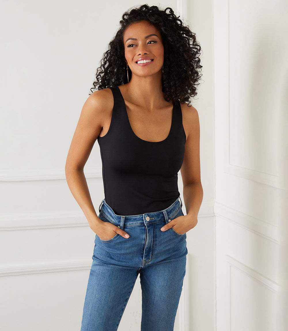 A woman with curly black hair wearing a black scoop-neck tank top and blue jeans, standing against a white paneled wall.