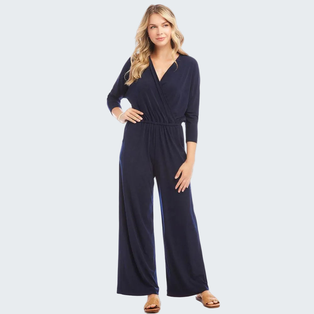 A woman modeling a navy blue wrap-style jumpsuit with three-quarter sleeves and wide legs, paired with tan flat sandals, against a plain white studio background.
