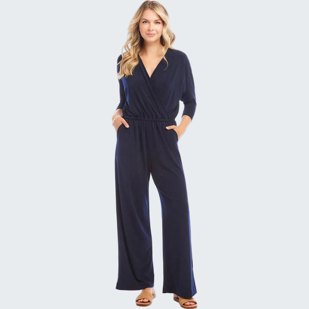 A woman modeling a navy blue wrap-style jumpsuit with three-quarter sleeves and wide legs, paired with tan slide sandals, against a plain white studio background.