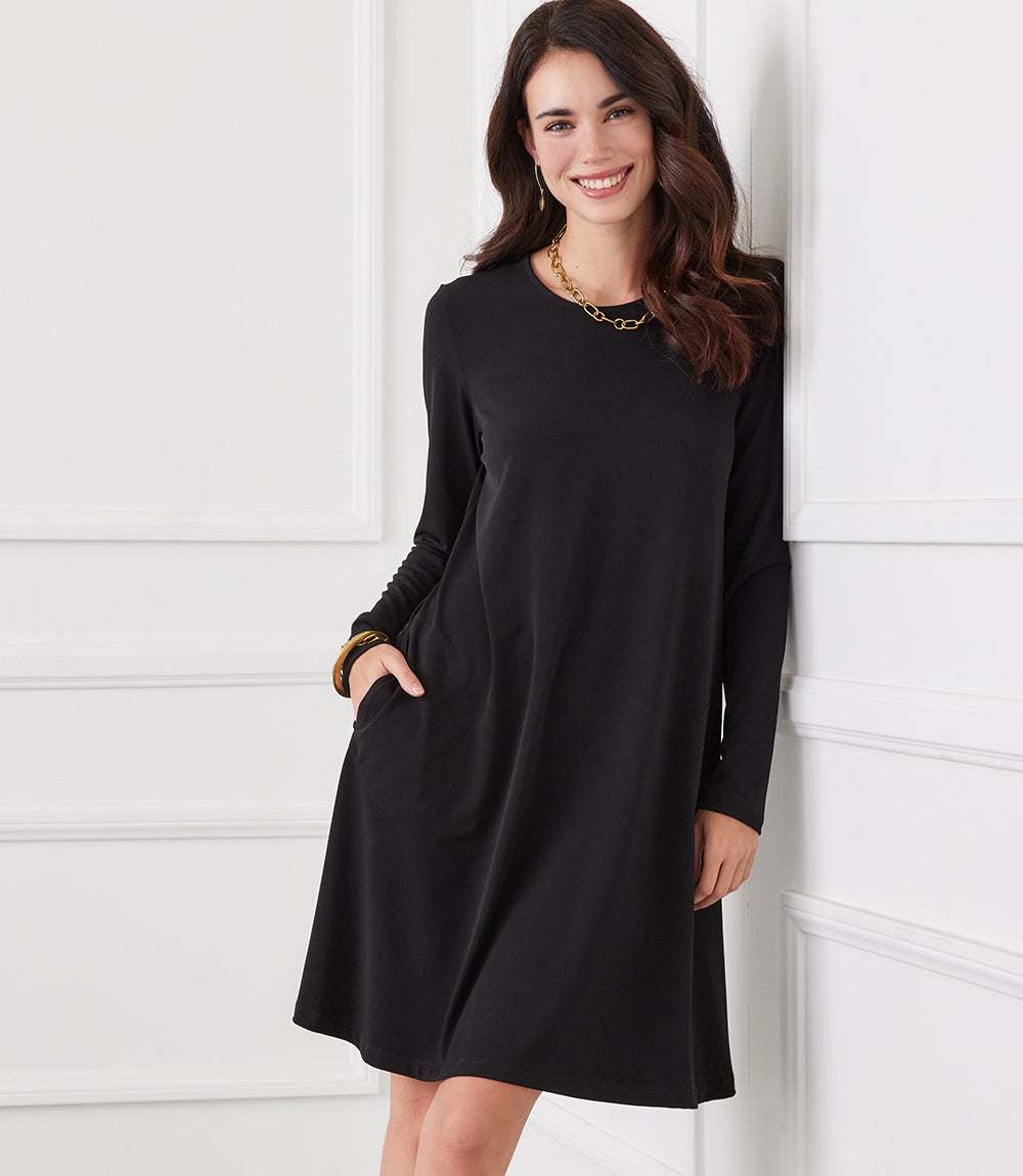 A woman wearing a black long-sleeve shift dress with side pockets, leaning against a white paneled wall.