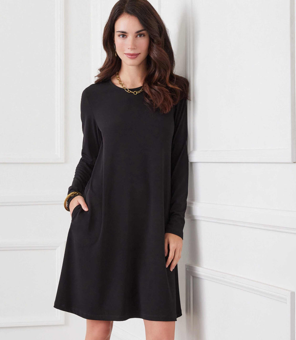 A woman wearing a black long-sleeve shift dress with side pockets, standing against a white paneled wall.
