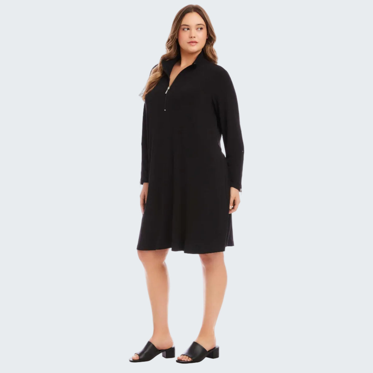 A woman modeling a black long-sleeve shift dress with a half-zip neckline, standing against a plain white studio background.