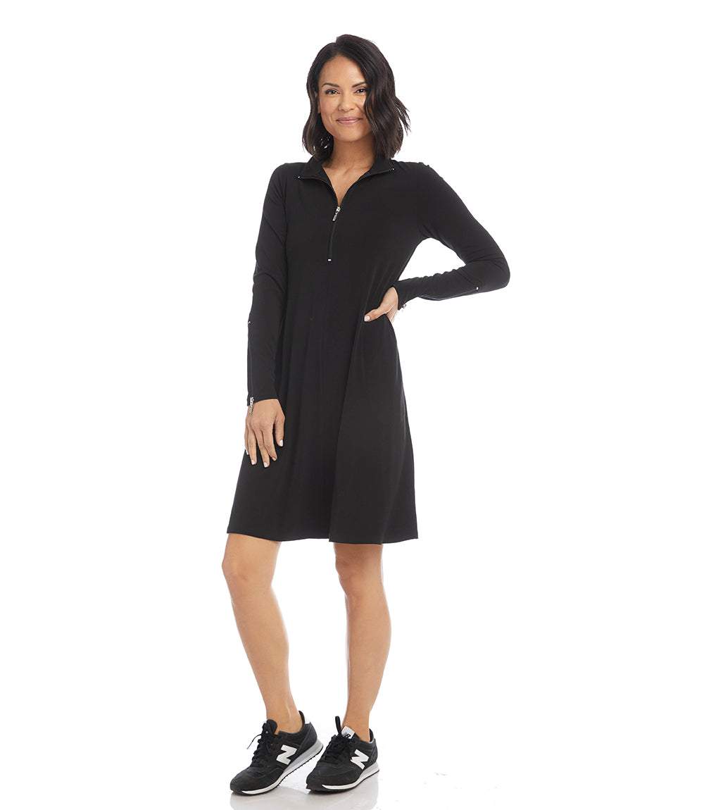 A woman modeling a black long-sleeve shift dress with a half-zip collar, paired with black sneakers, against a plain white studio background.