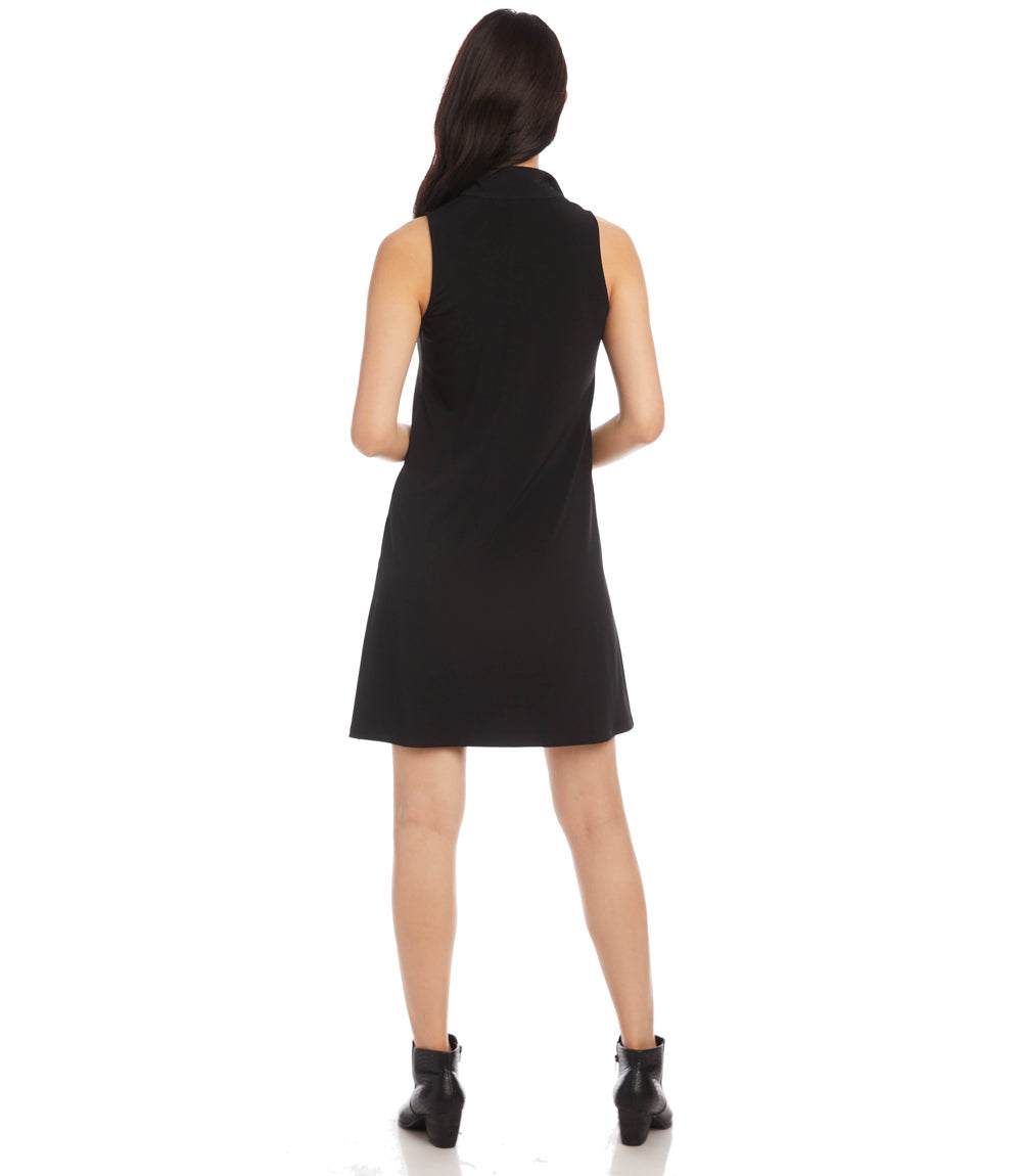 A woman viewed from the back wearing a sleeveless black shift dress and black ankle boots against a plain white studio background.