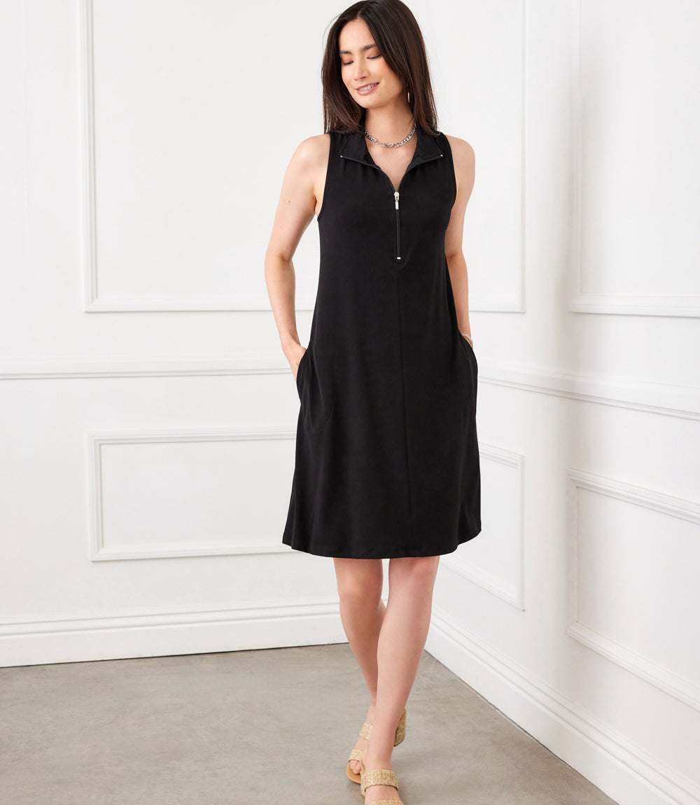 A woman modeling a sleeveless black dress with a half-zip collar and side pockets, standing in a white paneled room.