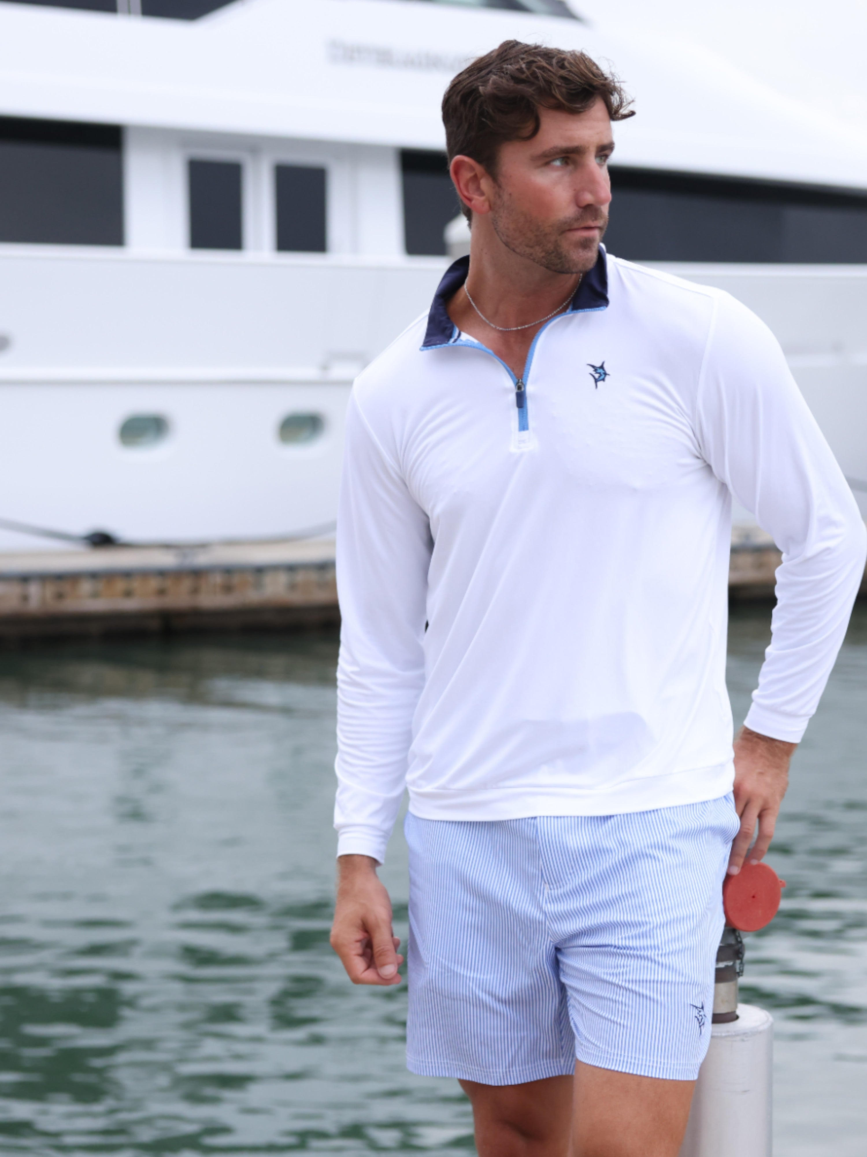 Man wearing a white long-sleeve shirt and light blue shorts standing by water with a boat in the background.