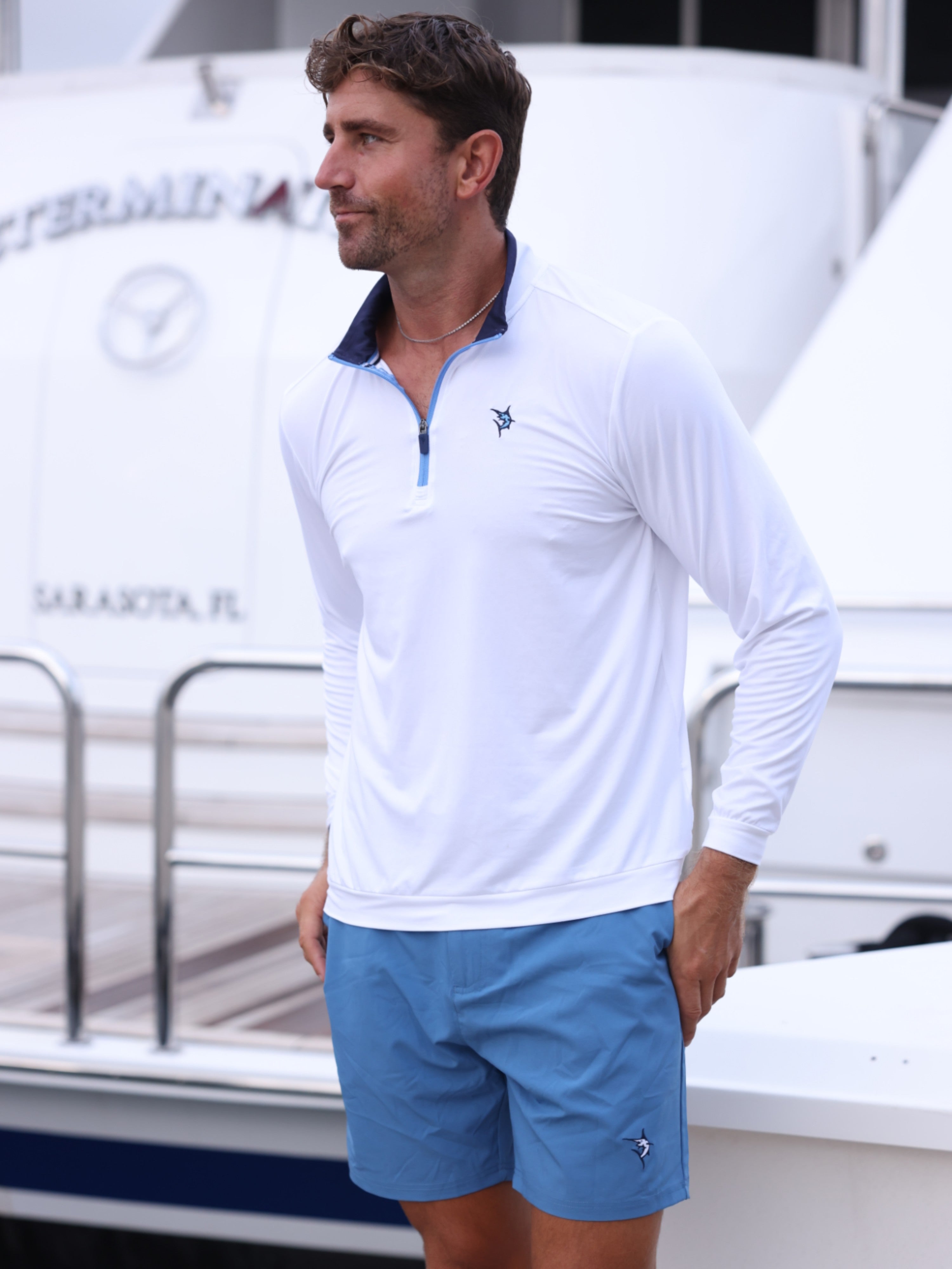 Man wearing a white long-sleeve shirt and blue shorts standing in front of a boat.
