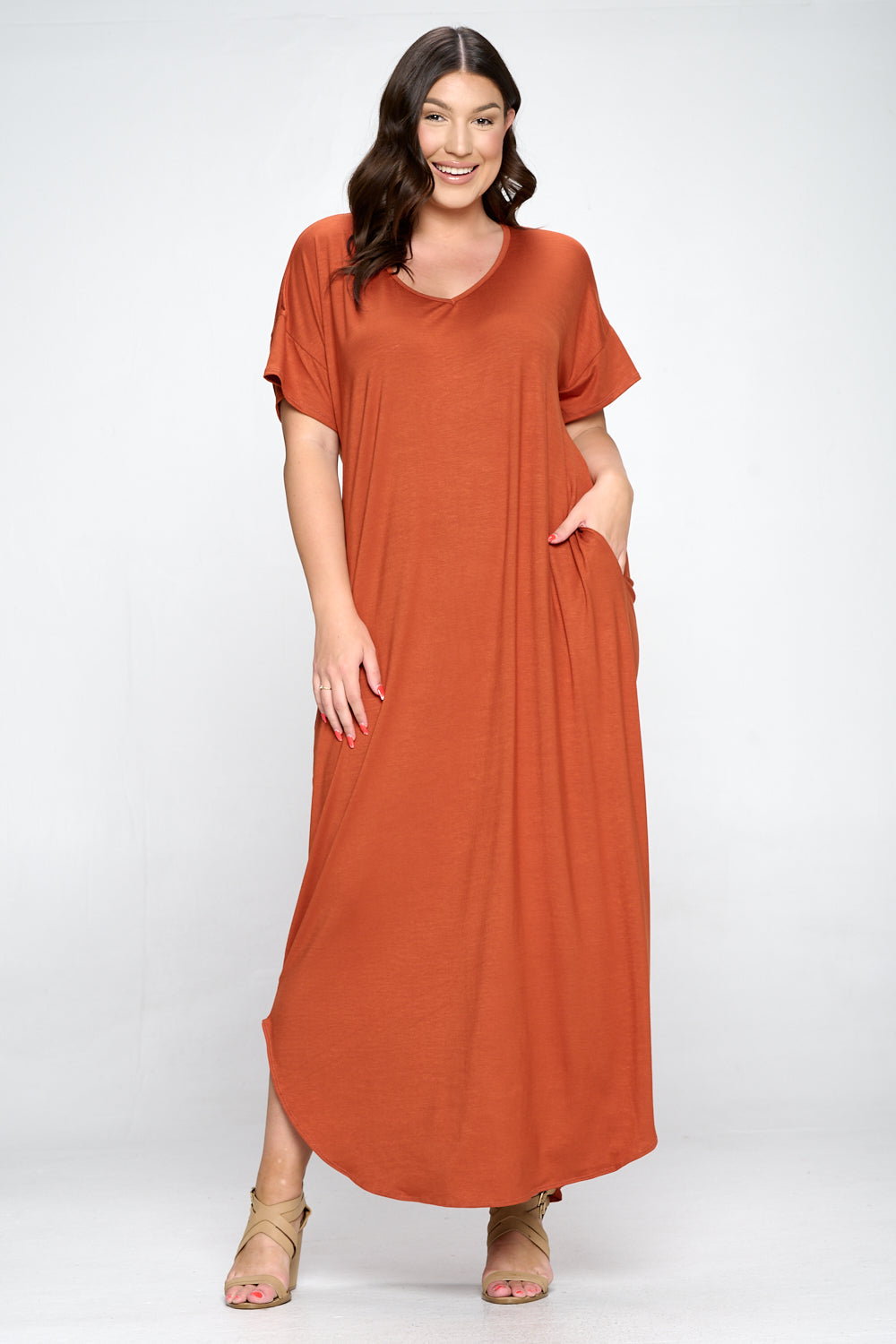 A woman modeling a loose, short-sleeve maxi dress in a warm rust orange color, standing against a plain light gray studio background.