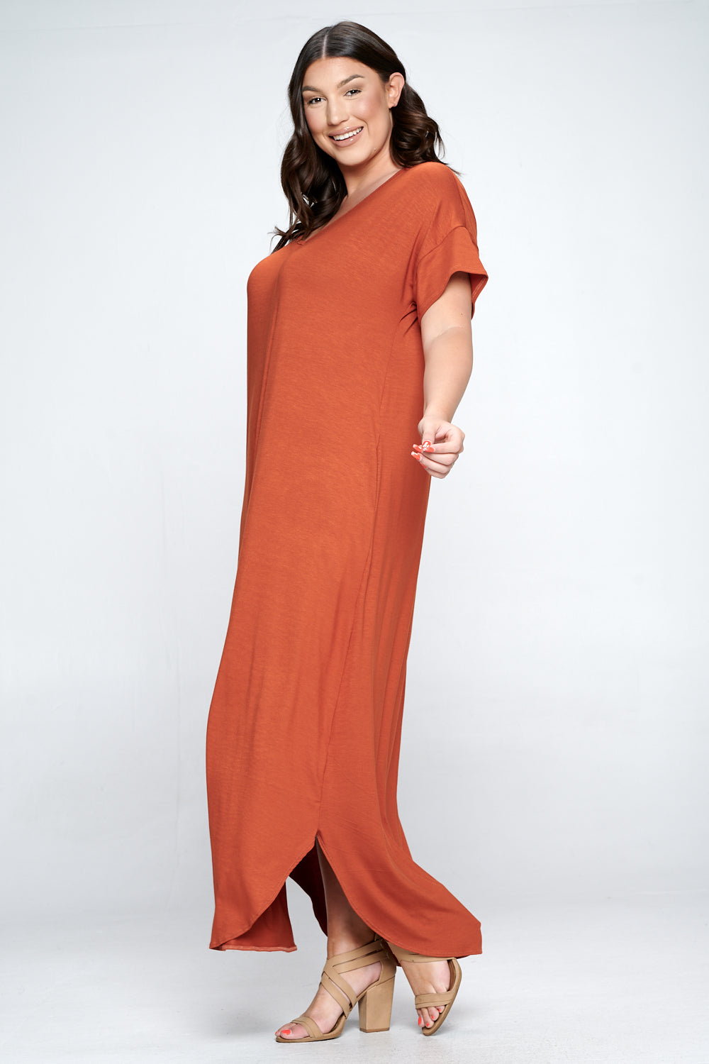 A woman modeling a rust-orange maxi dress with short flutter sleeves and a side slit, paired with beige heeled sandals, against a light gray studio background.