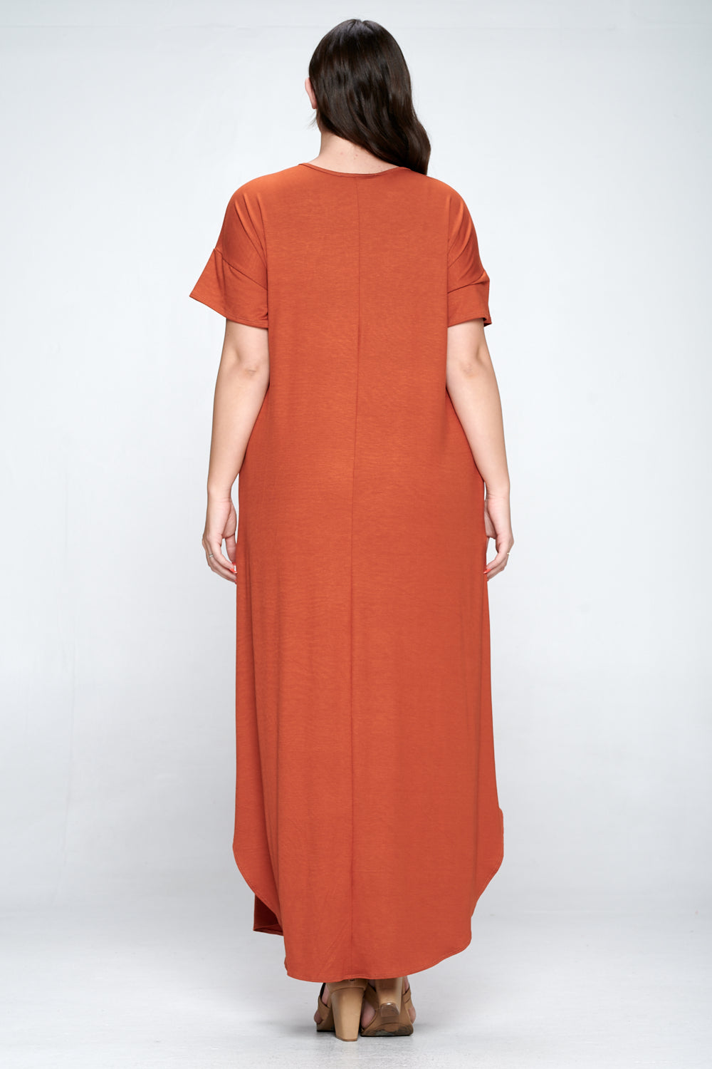 A woman viewed from the back wearing a loose, short-sleeve maxi dress in a warm rust orange color, paired with brown shoes, against a light gray studio background.