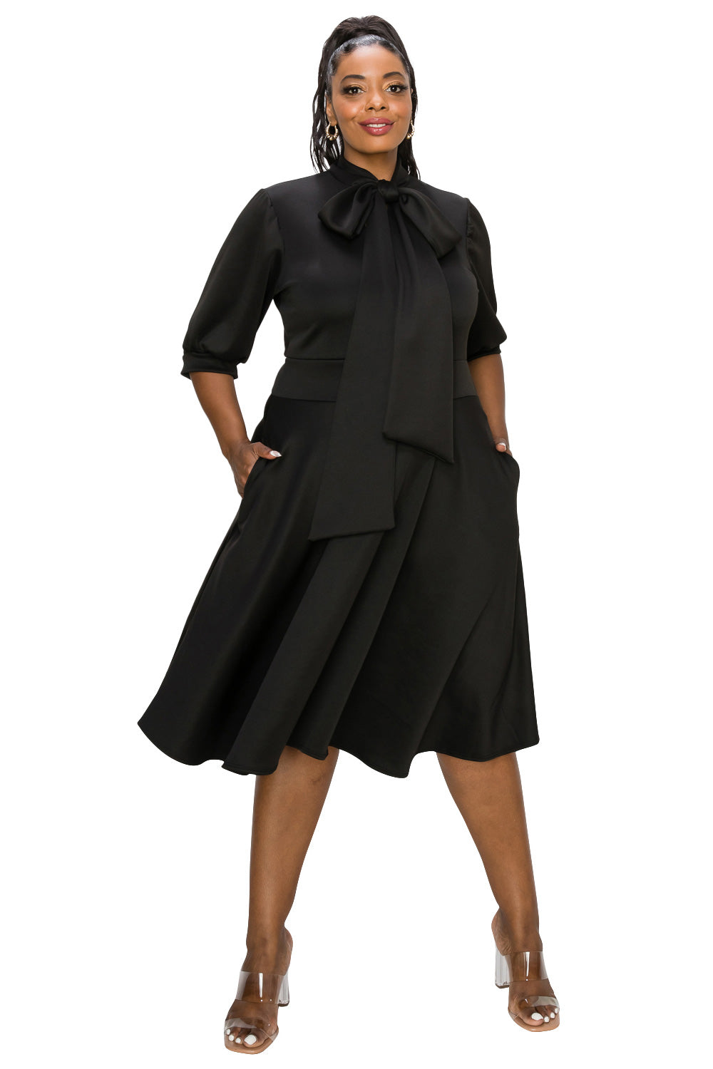 A woman modeling a black midi dress with a high neck, large bow detail at the collar, elbow-length sleeves, and a flared skirt, posed against a plain white background.
