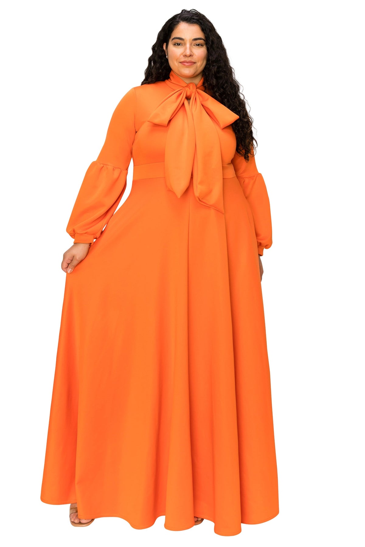 A woman modeling a bright orange long-sleeve maxi dress with a large bow tie at the high neckline, set against a plain white background.