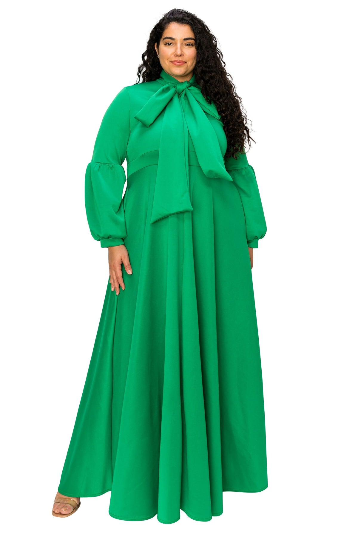 A woman modeling a long, flowing green dress with a large bow at the high neckline, long sleeves, and a full skirt, shown against a plain white background.