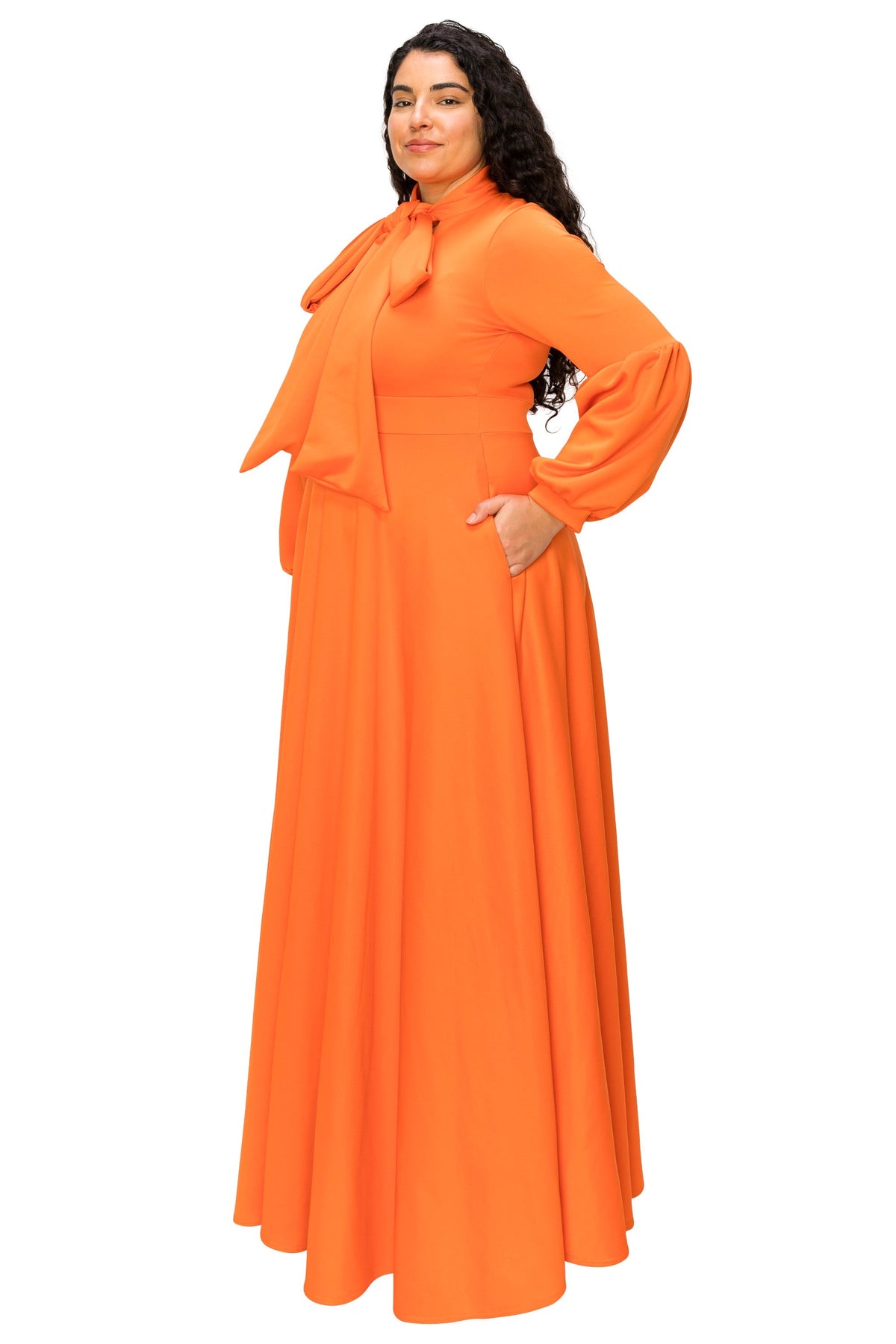 A woman modeling a bright orange long-sleeved maxi dress with a high neck, bow detail at the collar, and a flowing skirt, set against a plain white background.