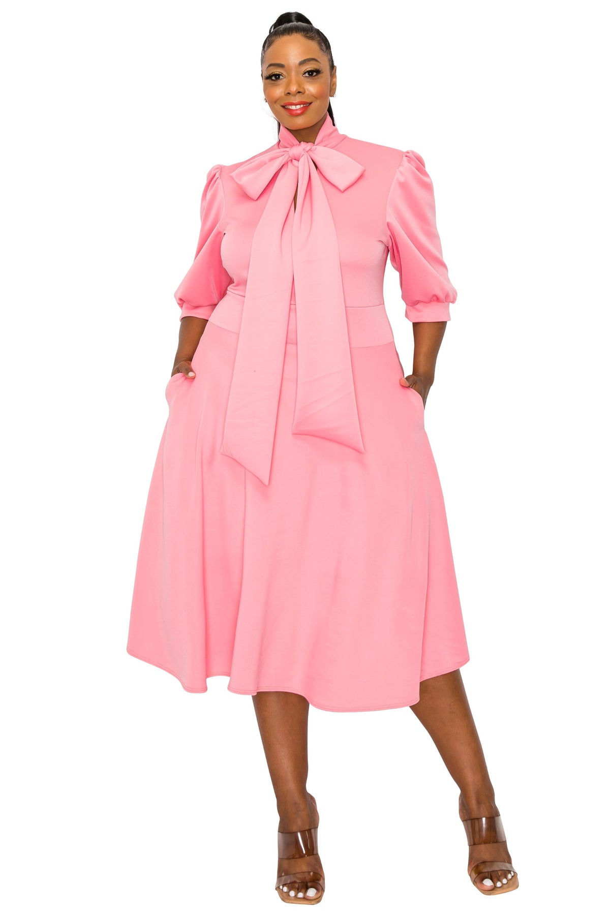 A woman modeling a pink midi dress with a large bow at the high neckline, puffed three-quarter sleeves, and a flared skirt, shown against a plain white background.