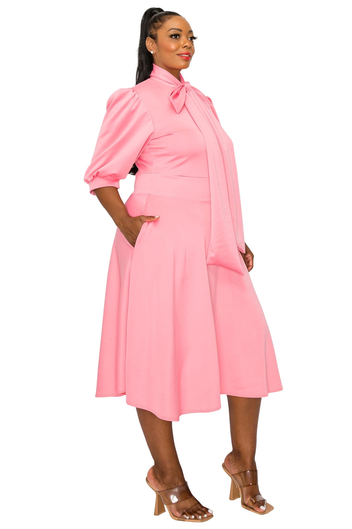 A woman modeling a light pink midi dress with puff sleeves, a high collar with a bow, and a flared skirt, paired with brown heeled sandals against a plain white background.