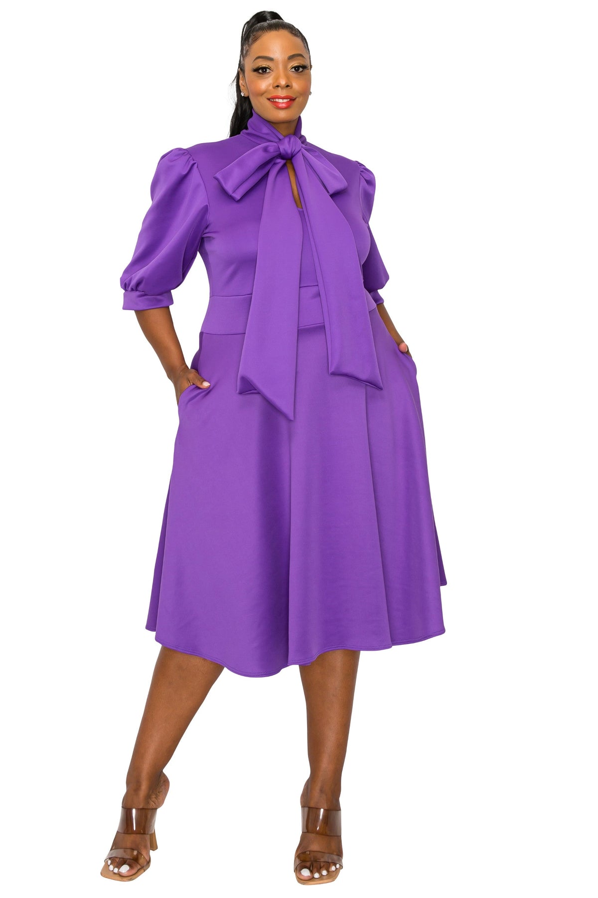 A woman modeling a bright purple midi dress with puff sleeves, a large bow tie neckline, and side pockets, paired with brown heeled sandals against a plain white background.