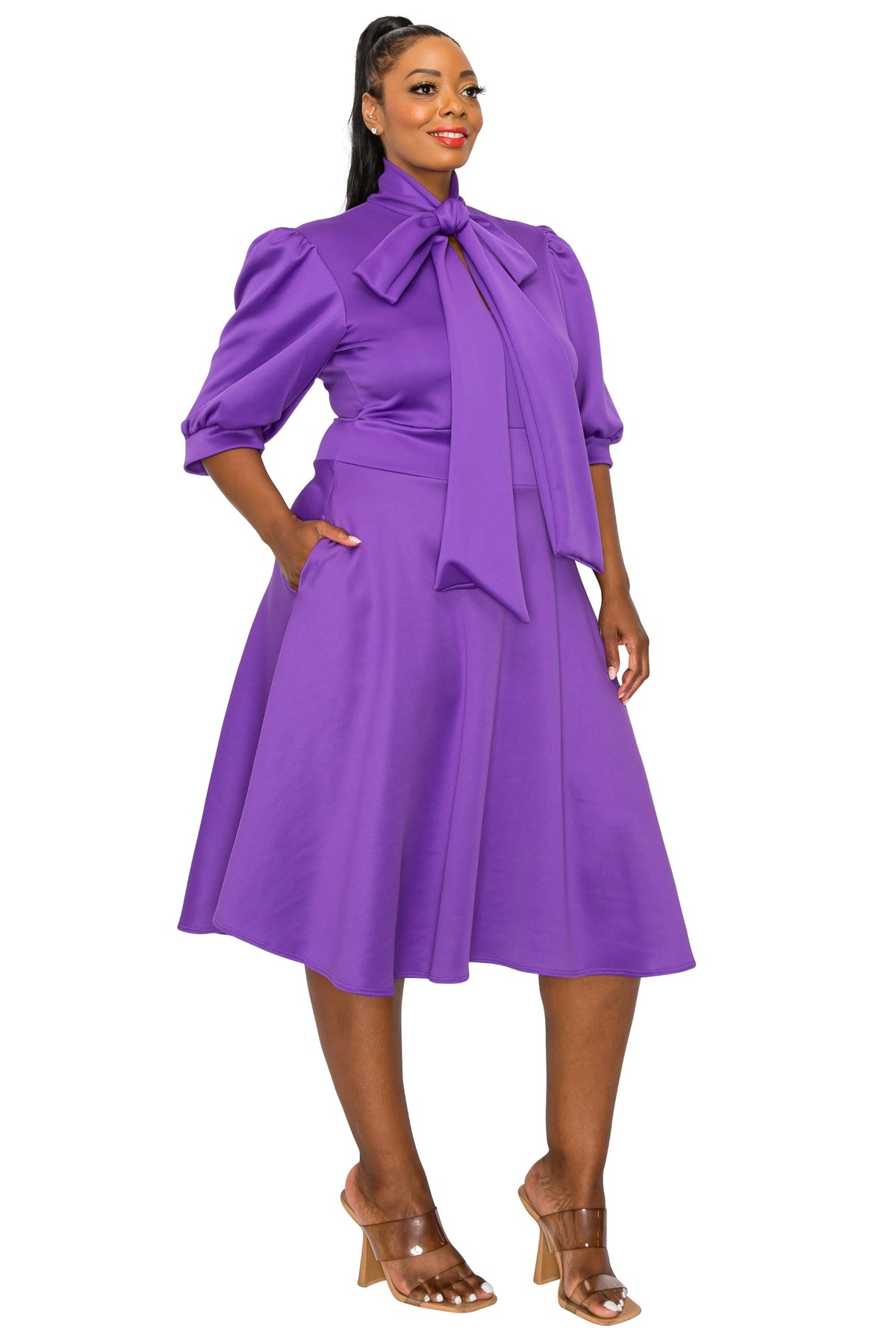 A woman modeling a bright purple midi dress with a high neck, large bow at the collar, puffed three-quarter sleeves, a fitted waist, and a flared skirt, paired with clear-strap heeled sandals against a plain white background.