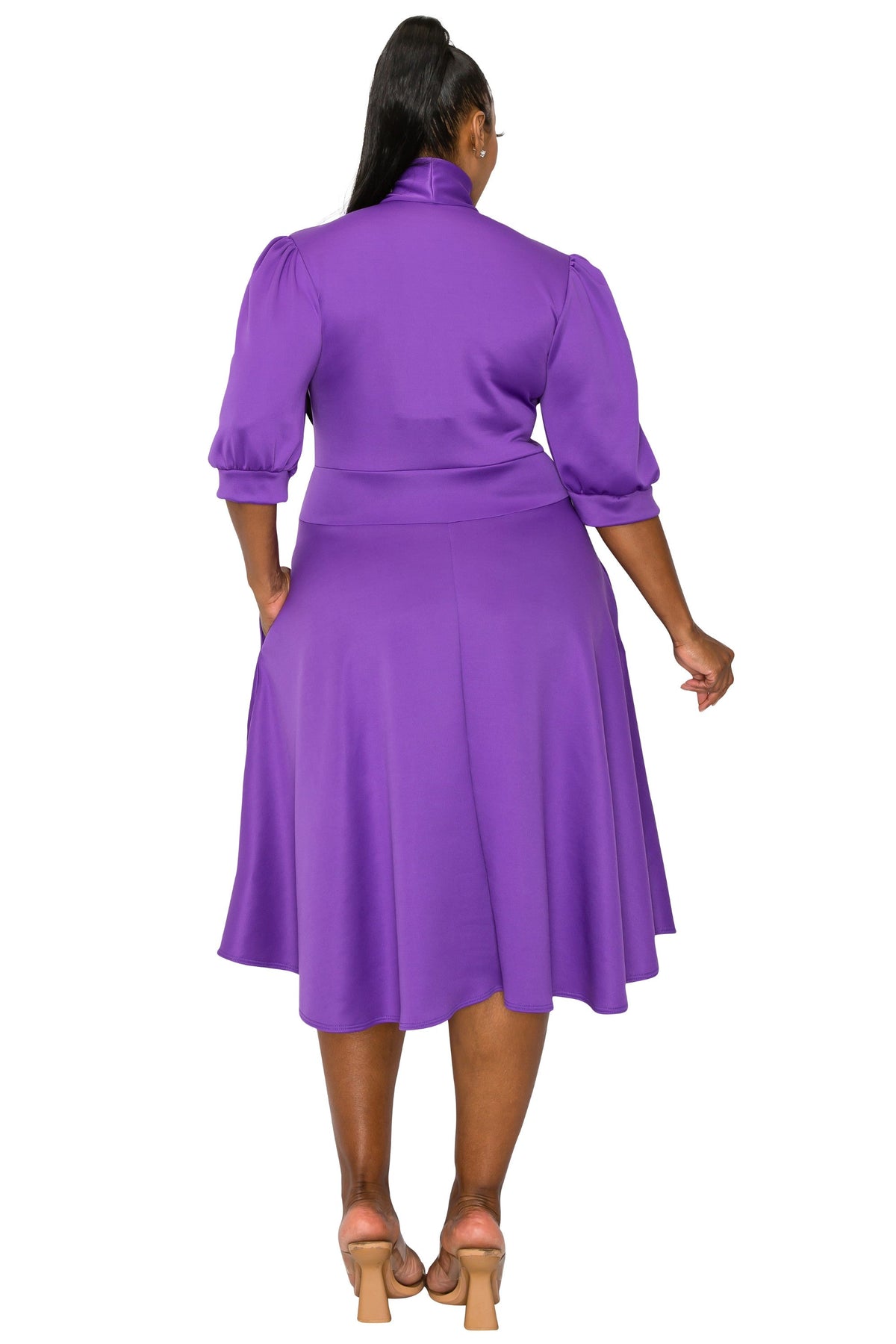 A woman viewed from the back wearing a purple midi dress with puff sleeves and a flared skirt, paired with tan high-heeled sandals, against a plain white background.