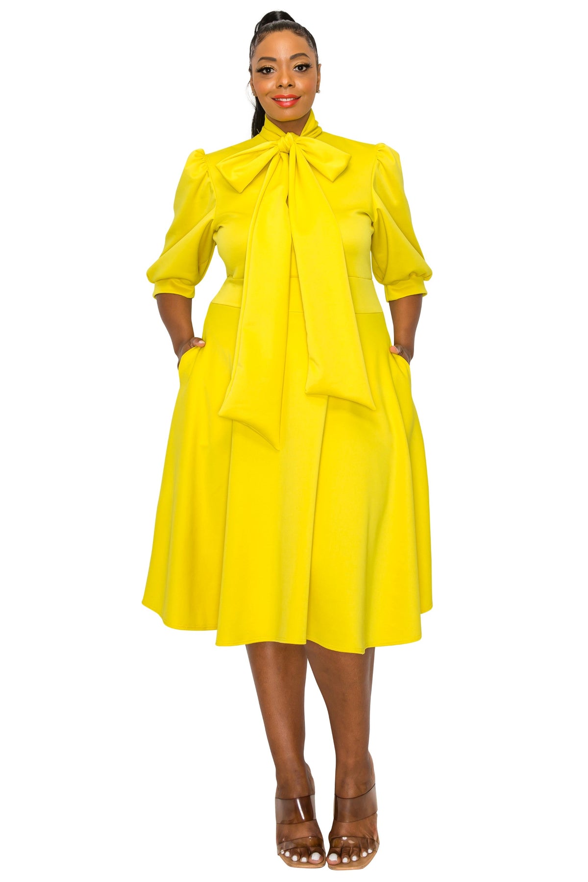 A woman modeling a bright yellow midi dress with a high neck, large bow at the collar, three-quarter sleeves, and side pockets, paired with brown strappy heels against a plain white background.