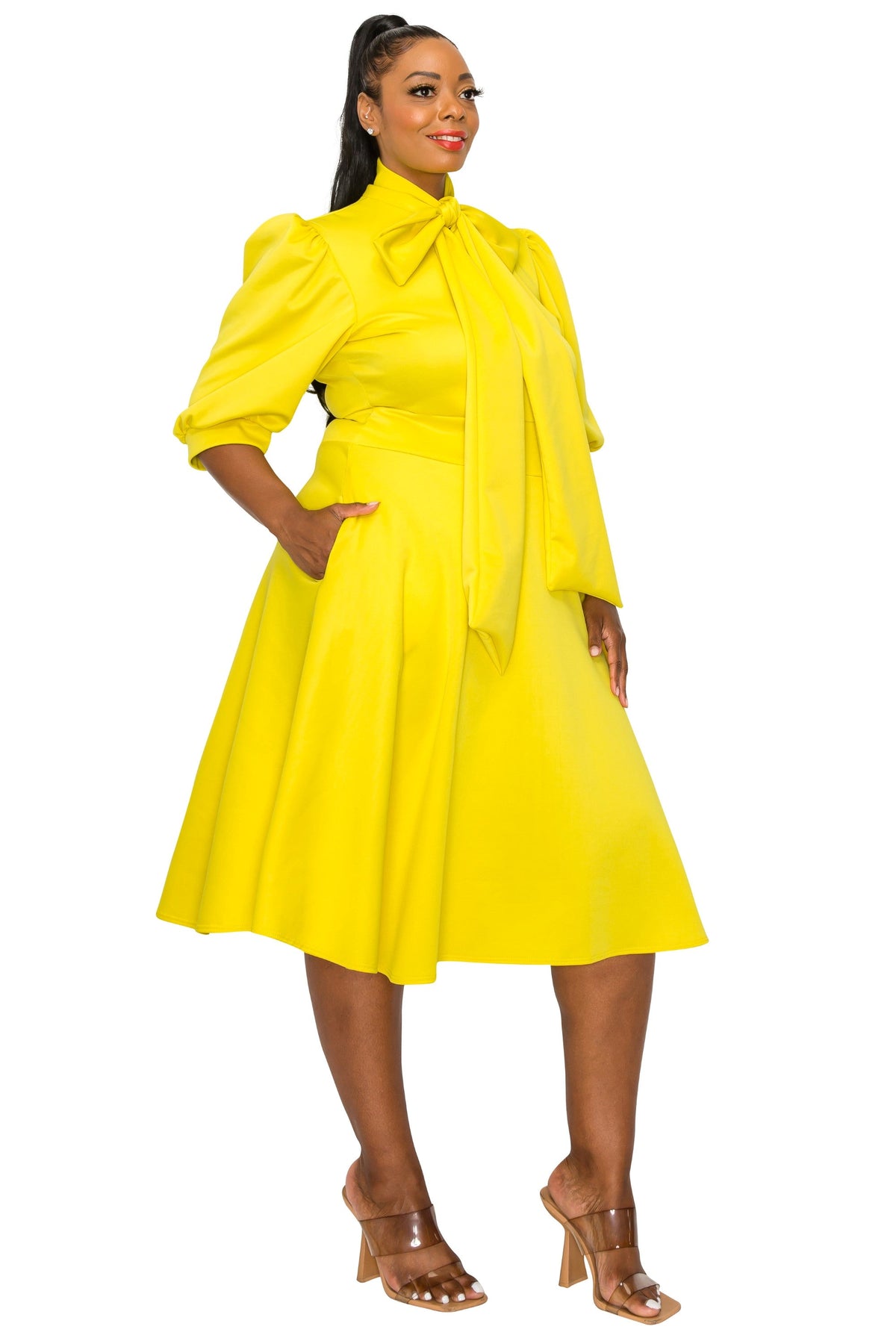 A woman modeling a bright yellow midi dress with puffed elbow-length sleeves, a high collar with a large bow tie-front neckline, a fitted waist, and a flared skirt. She is wearing clear high-heeled sandals against a plain white studio background.