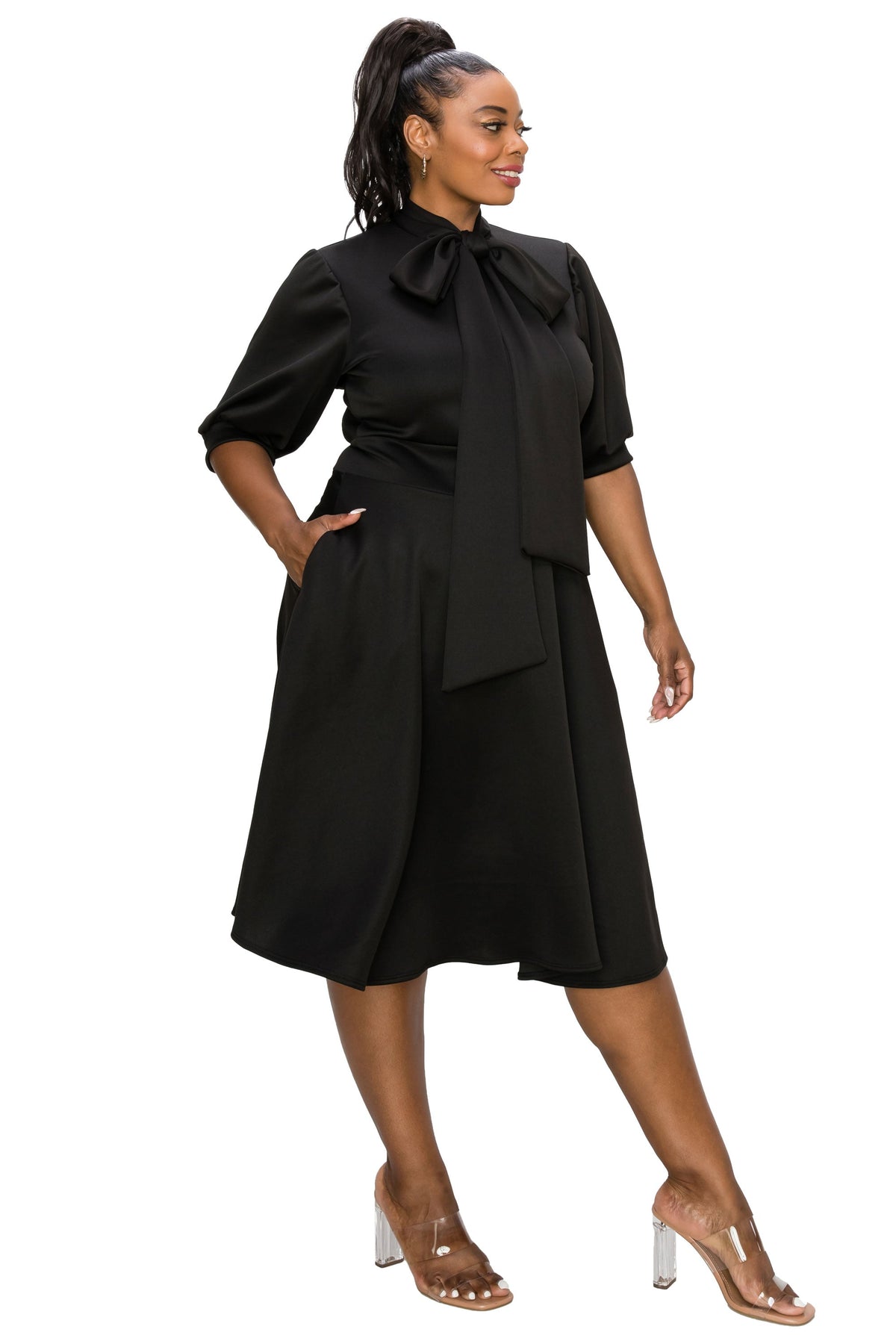 A woman modeling a black midi dress with a high neck, large bow at the collar, elbow-length puff sleeves, side pockets, and a draped waist detail, paired with clear high-heel sandals against a plain white background.