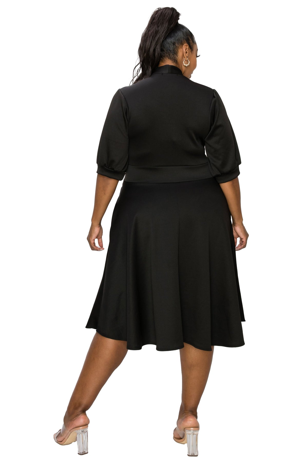 A woman shown from the back wearing a black midi dress with three-quarter sleeves and a flared skirt, paired with clear high-heeled sandals, against a plain white background.