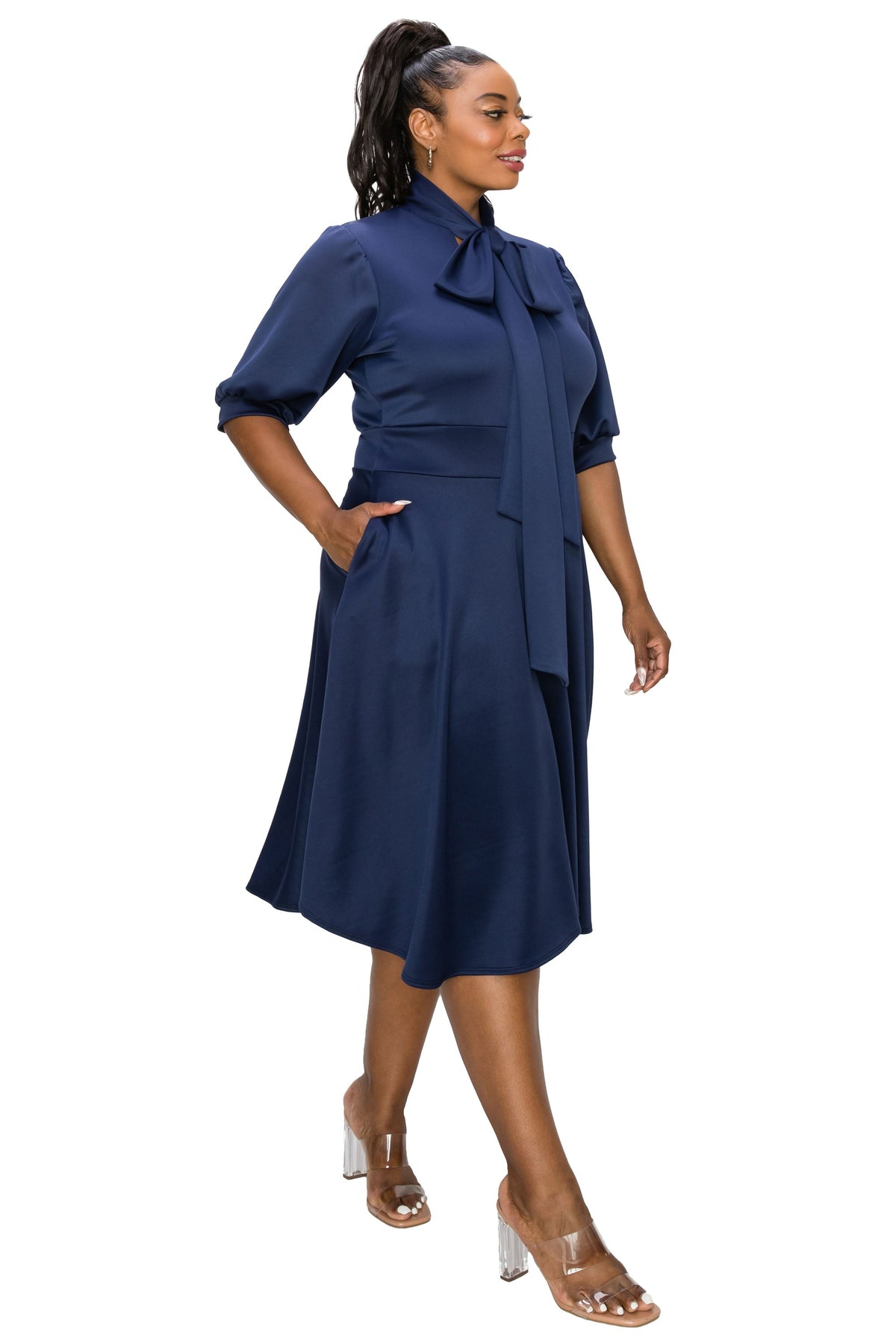 A woman modeling a navy blue midi dress with a high neck, bow detail at the collar, elbow-length sleeves, side pockets, and a draped waist tie, paired with clear high-heeled sandals against a plain white background.