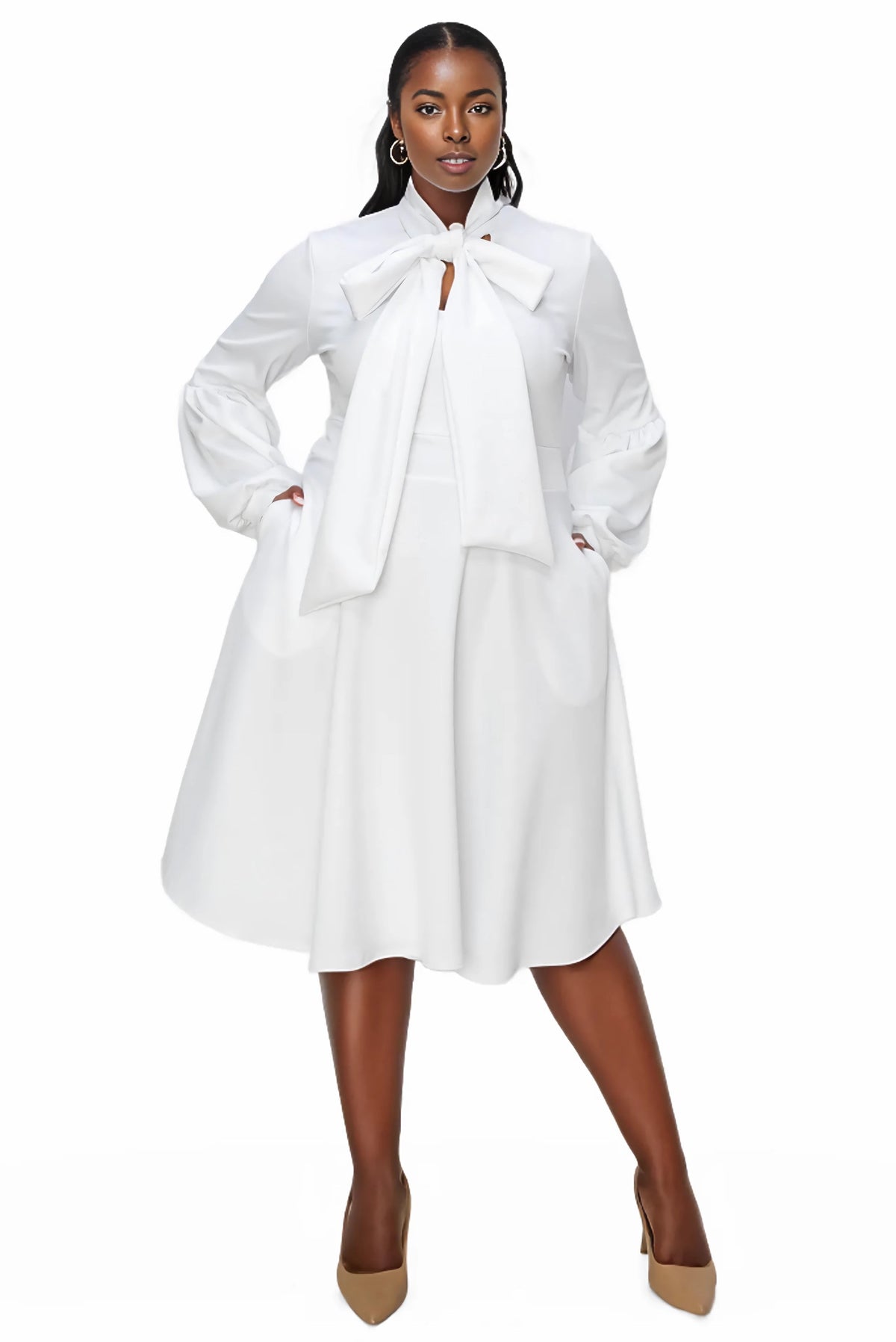 A woman modeling a white long-sleeve midi dress with a large bow at the collar, side pockets, and a flared skirt, paired with tan pointed-toe heels against a plain white background.