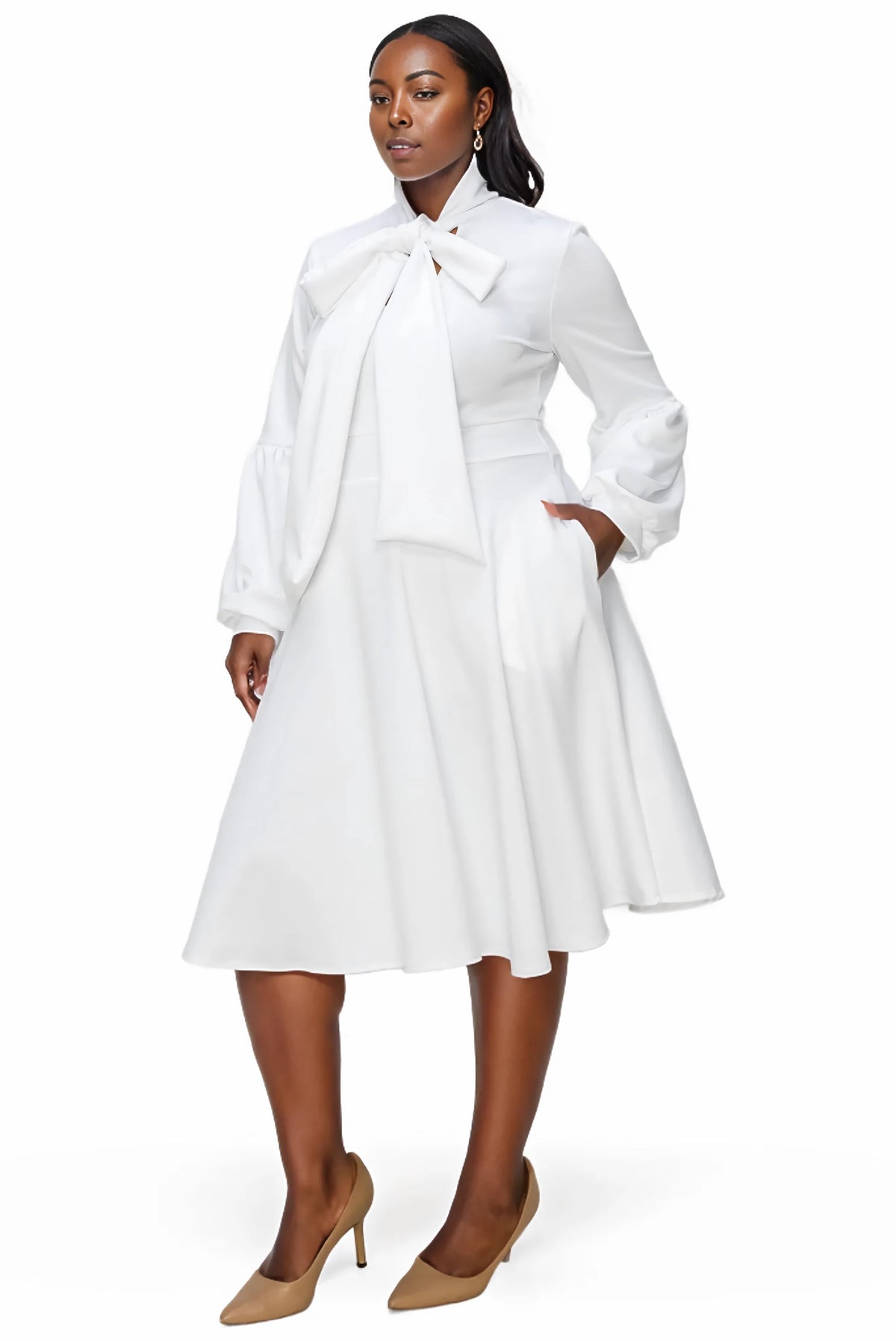 A woman modeling a white long-sleeve midi dress with a large bow at the collar, side pockets, and a flared skirt, paired with tan pointed-toe heels against a plain white studio background.