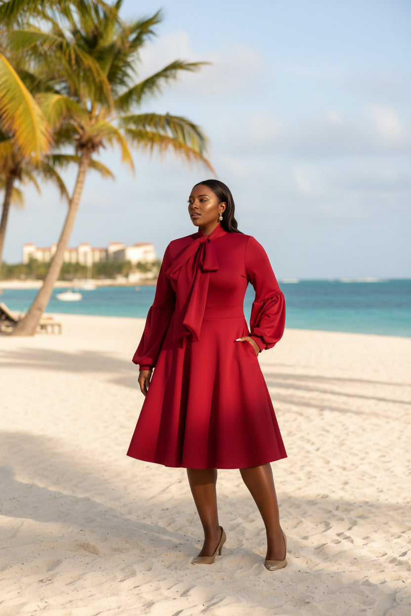A woman modeling a long-sleeve midi dress in deep burgundy red, featuring a large bow at the high neckline, puff sleeves, and a flared skirt, paired with tan heels against a plain white studio background.