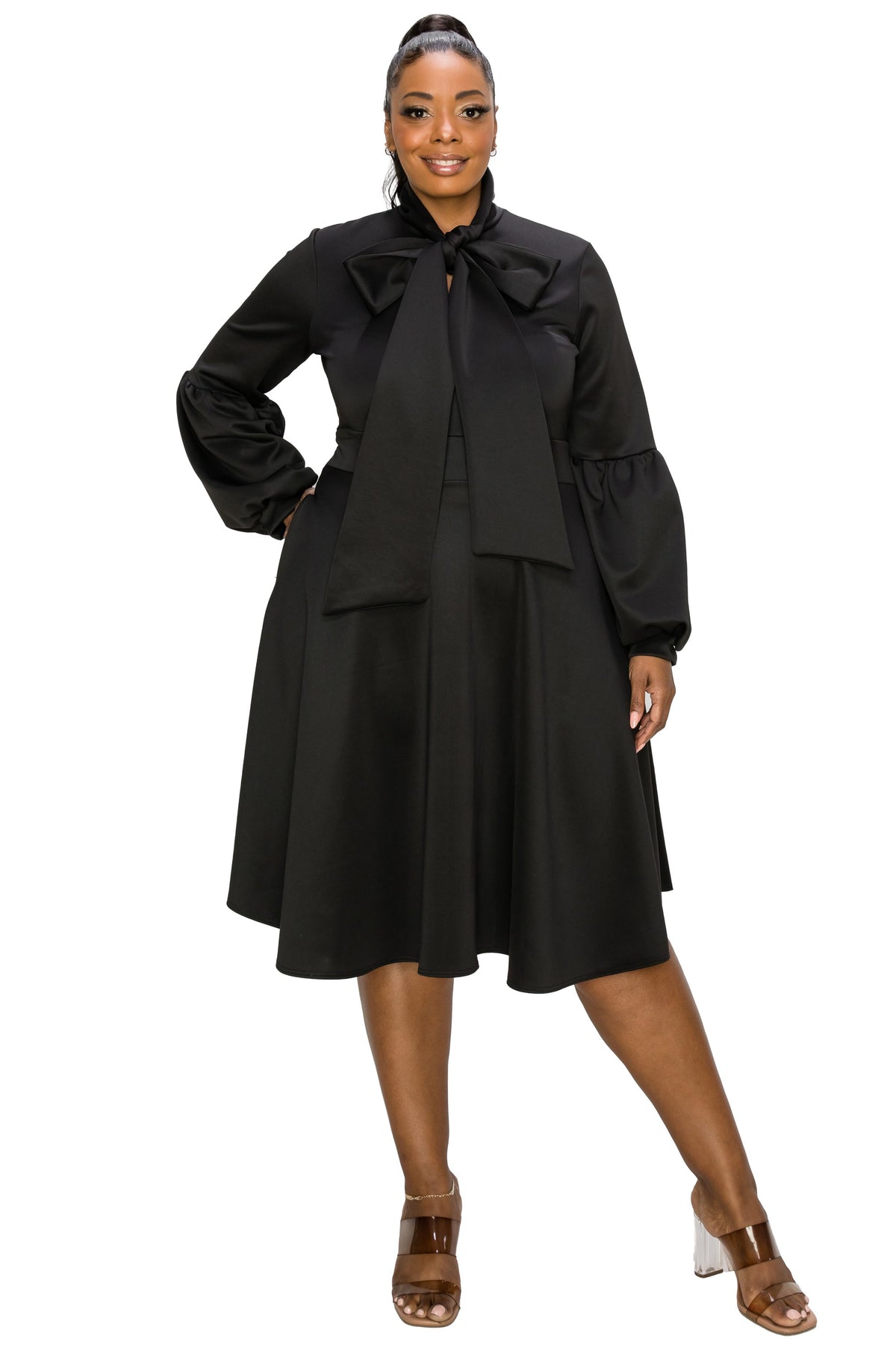 A woman modeling a black long-sleeve midi dress with a large bow at the collar, puff sleeves, and a flared skirt, paired with brown heeled sandals against a plain white background.