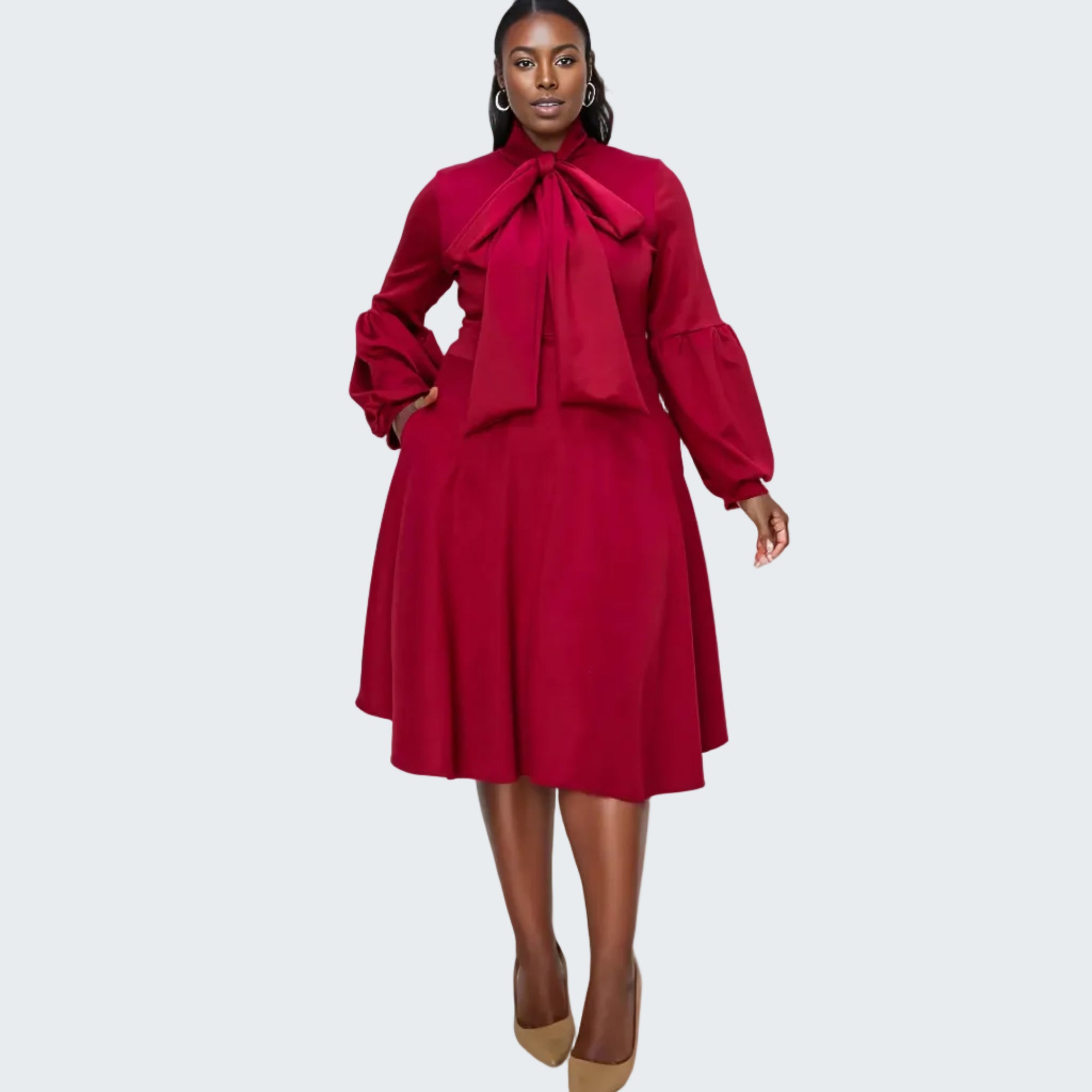 A woman modeling a long-sleeve midi dress in deep red with a large bow tie at the collar, puffed balloon sleeves, and a flared skirt, paired with tan pointed-toe heels against a plain white background.