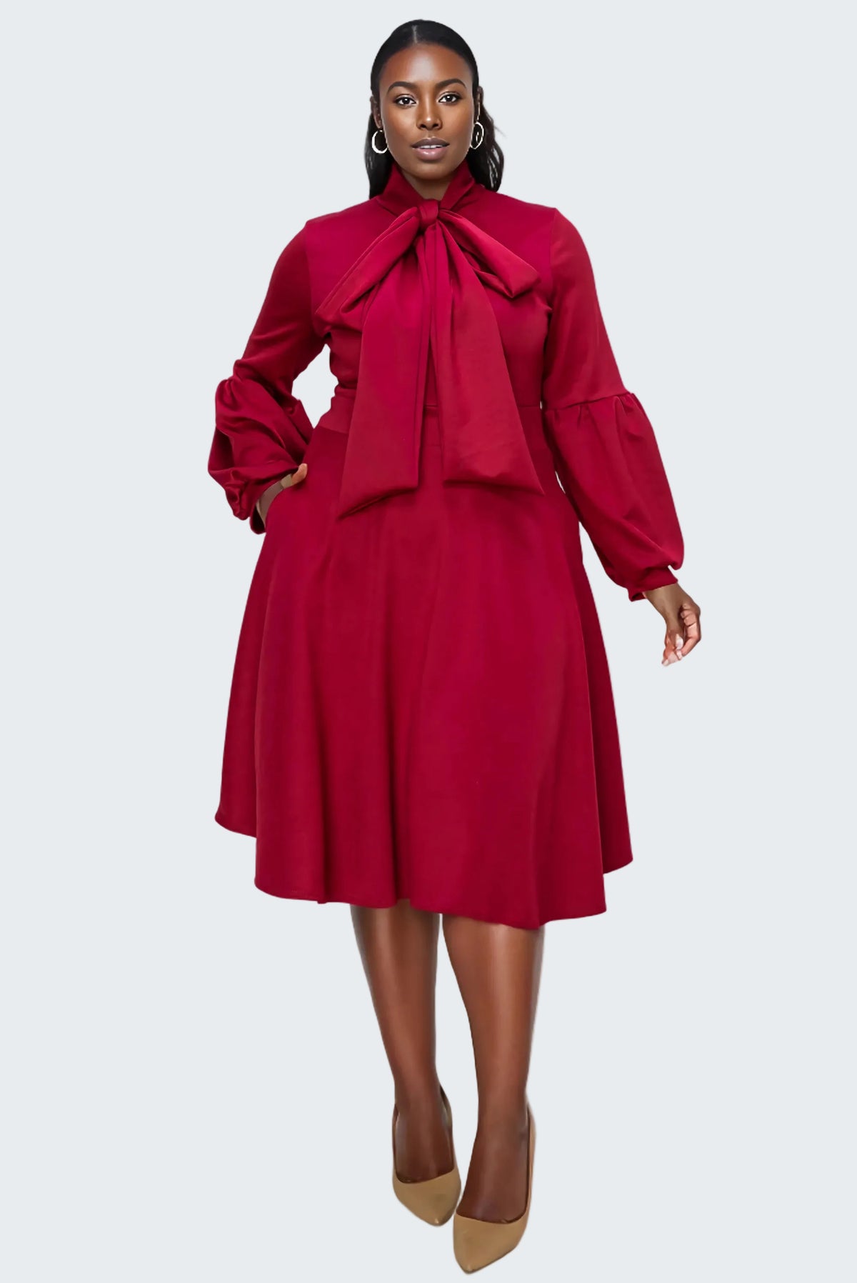 A woman modeling a long-sleeve midi dress in deep red with a large bow tie at the collar, puffed balloon sleeves, and a flared skirt, paired with tan pointed-toe heels against a plain white background.
